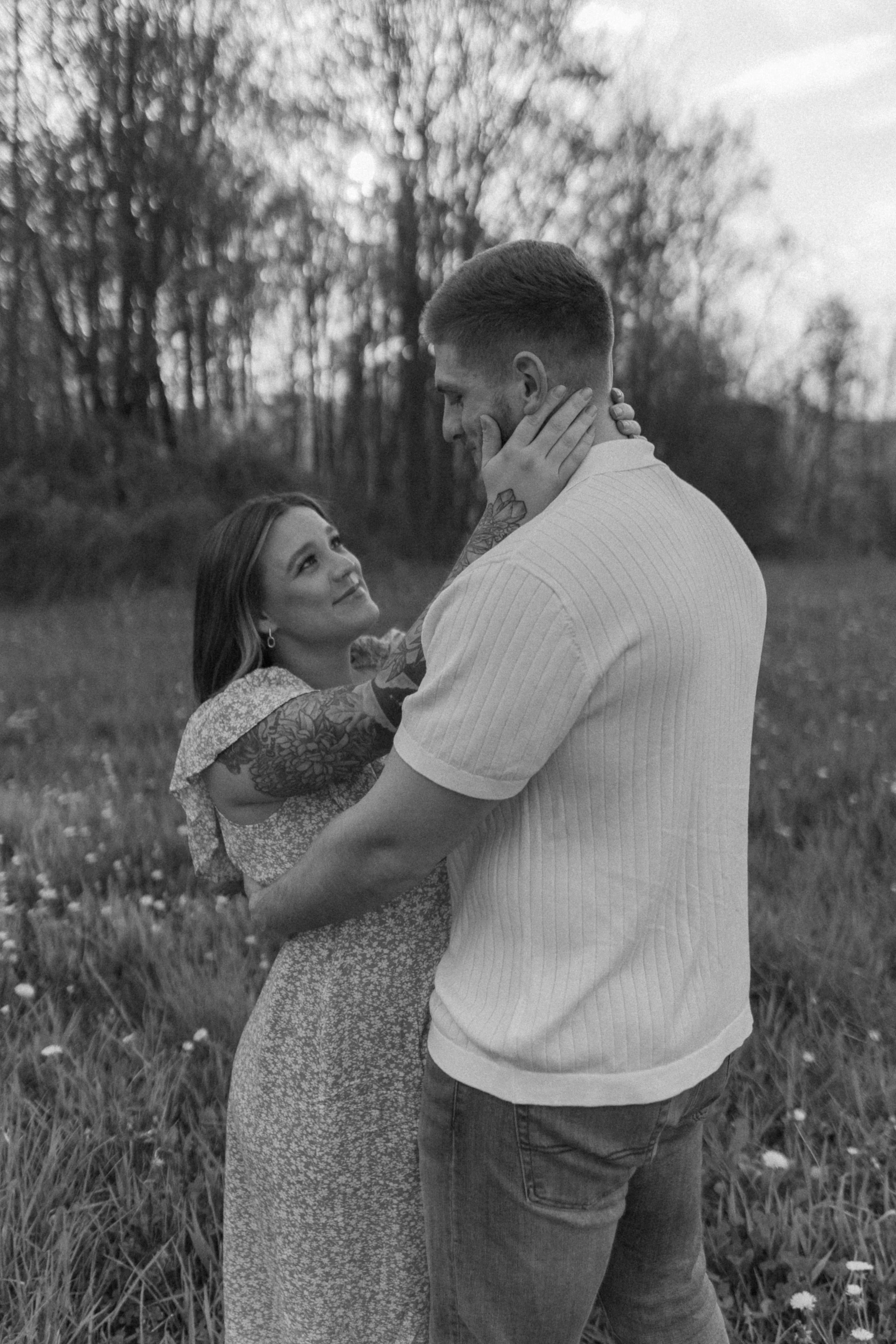 A black-and-white photo of a couple standing outdoors in a field with trees in the background. The woman is holding the man's face with both hands, and they are looking into each other's eyes, smiling.