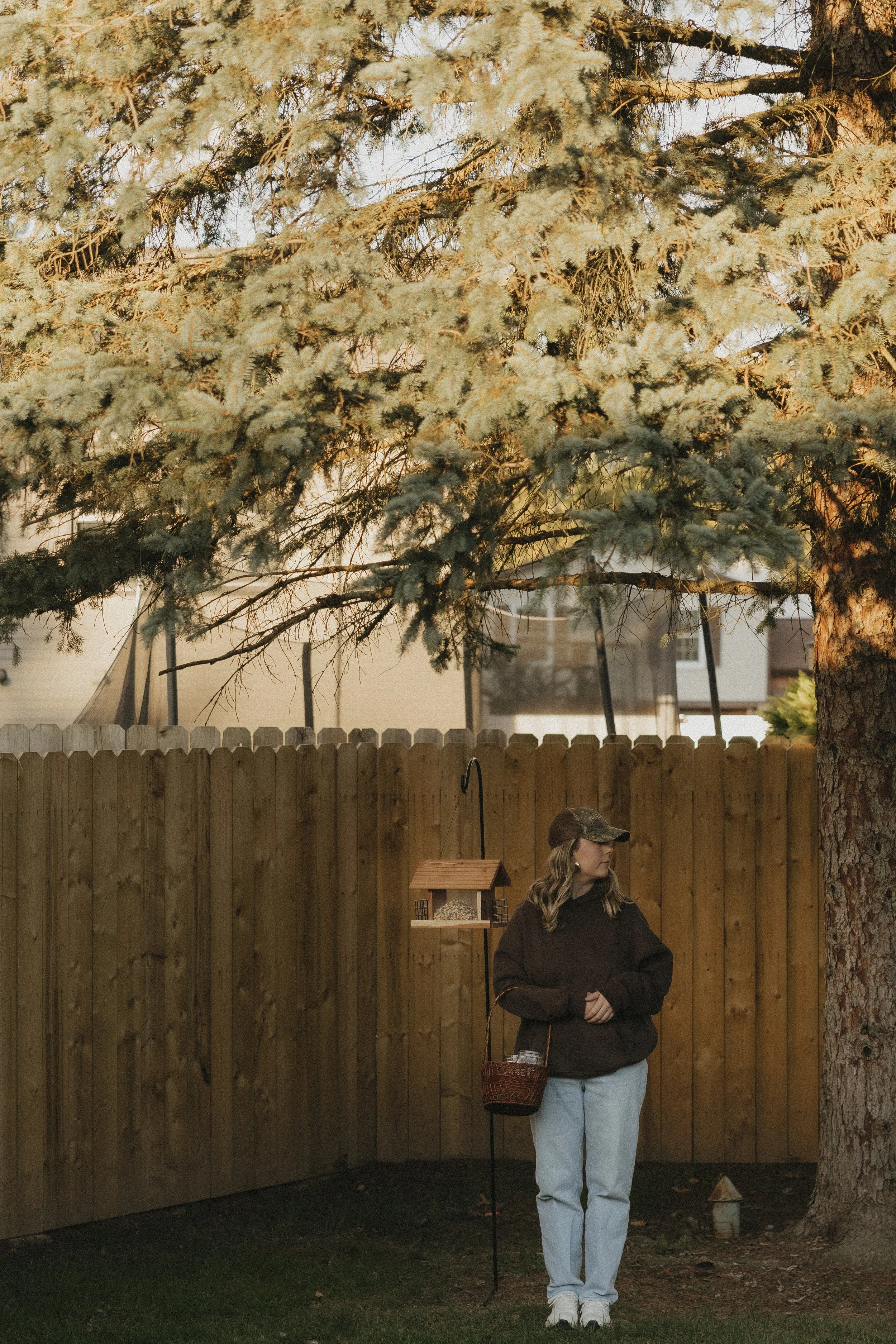 A woman wearing a dark hoodie and light-colored pants standing outdoors near a wooden fence. She has a basket over her shoulder and a camouflage hat, with a bird feeder hanging on a pole beside her. Tall pine trees are in the background, and it appears to be late afternoon or early evening.