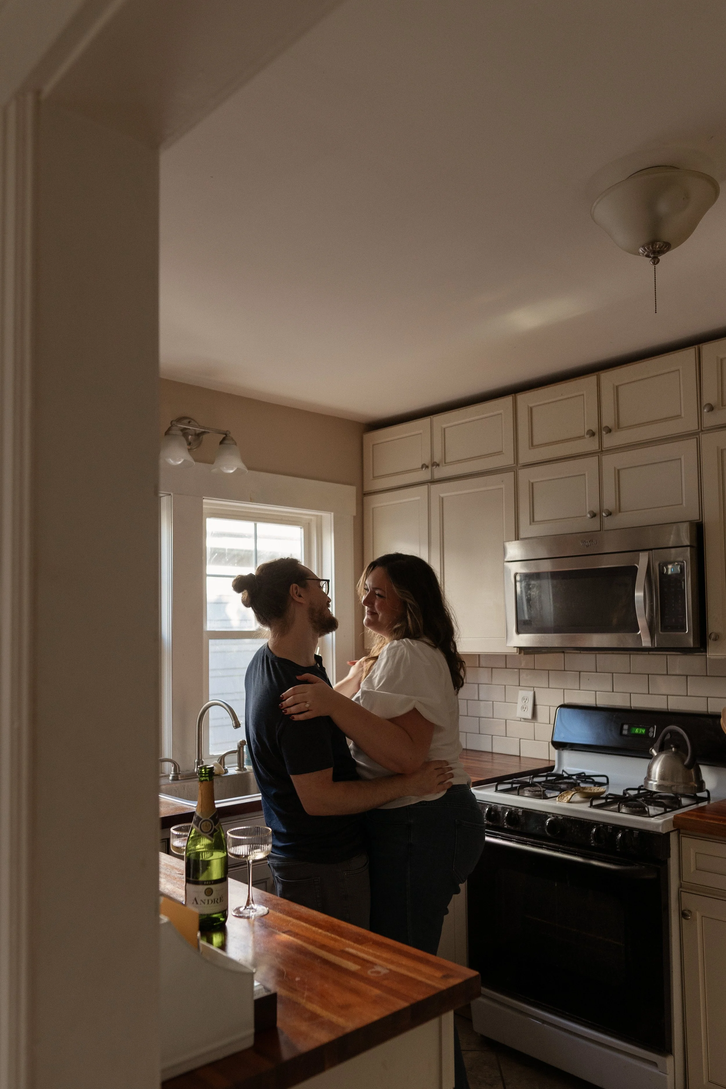 A couple in a kitchen, embracing and smiling at each other, with wine glasses and a bottle of champagne on the counter.