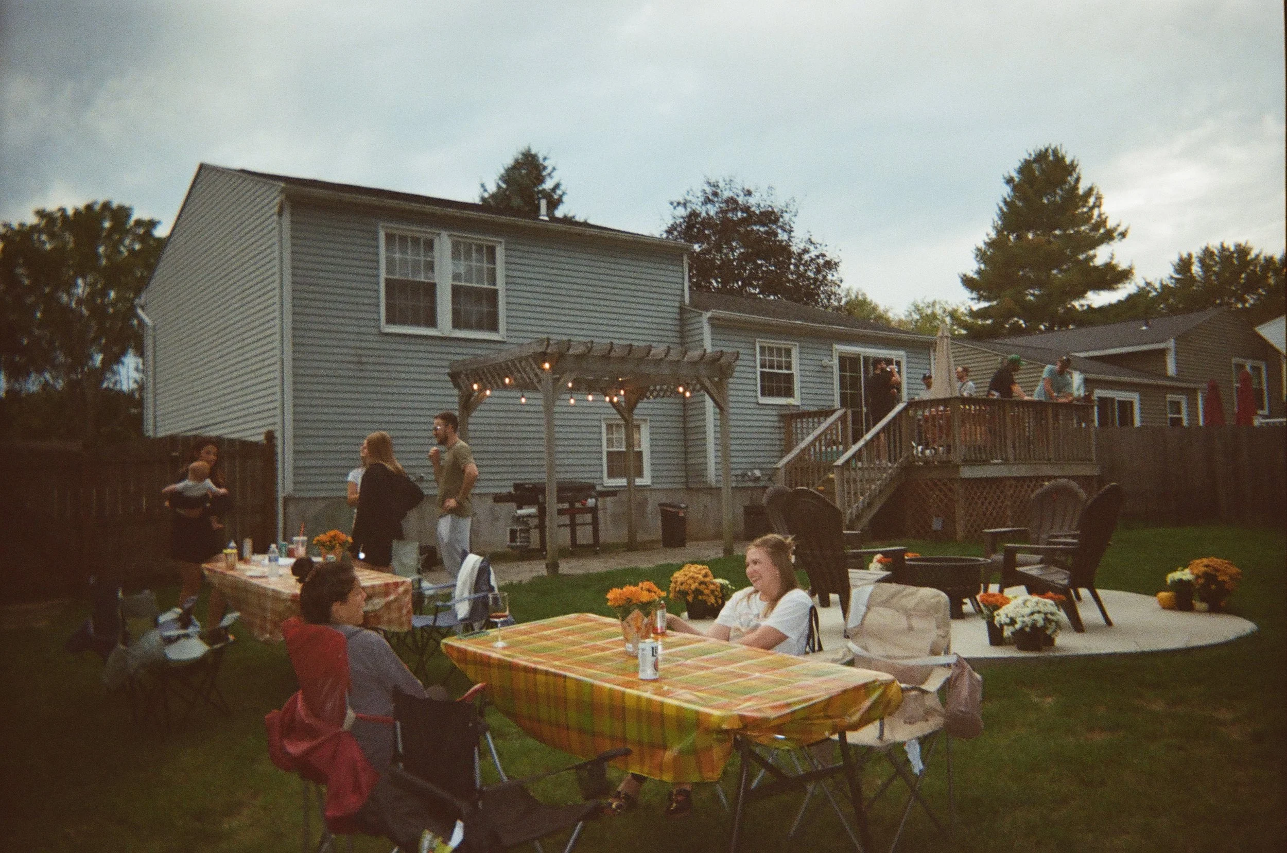 People gathered outdoors in a backyard, some sitting at tables with yellow and orange checkered tablecloths, others standing and talking near a house with a deck, a pergola with string lights, and potted flowers. The scene appears to be a social gathering or party during the evening.