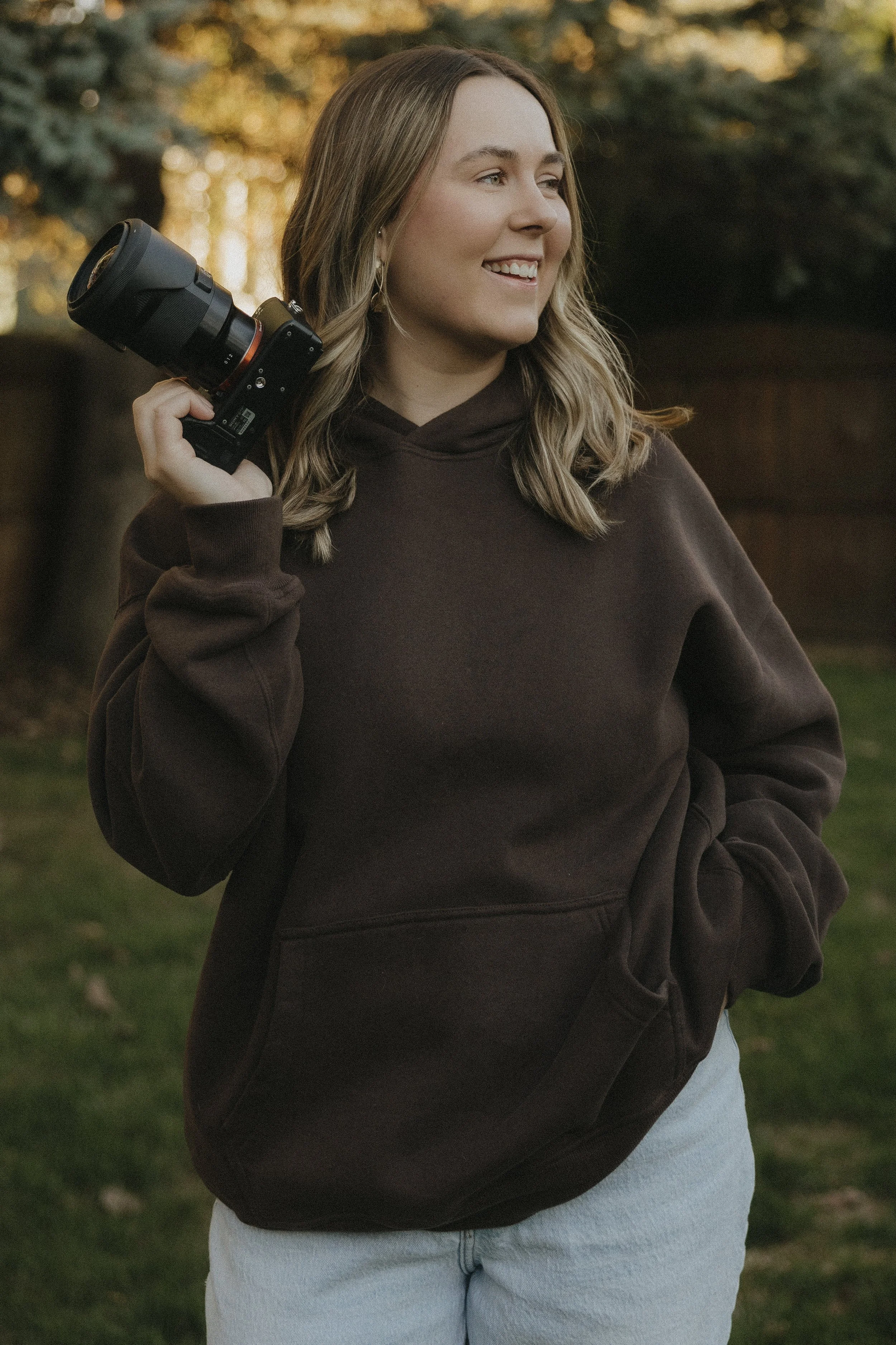 Young woman outdoors holding a camera on her shoulder, smile, brown hoodie, light-colored jeans, trees in the background.