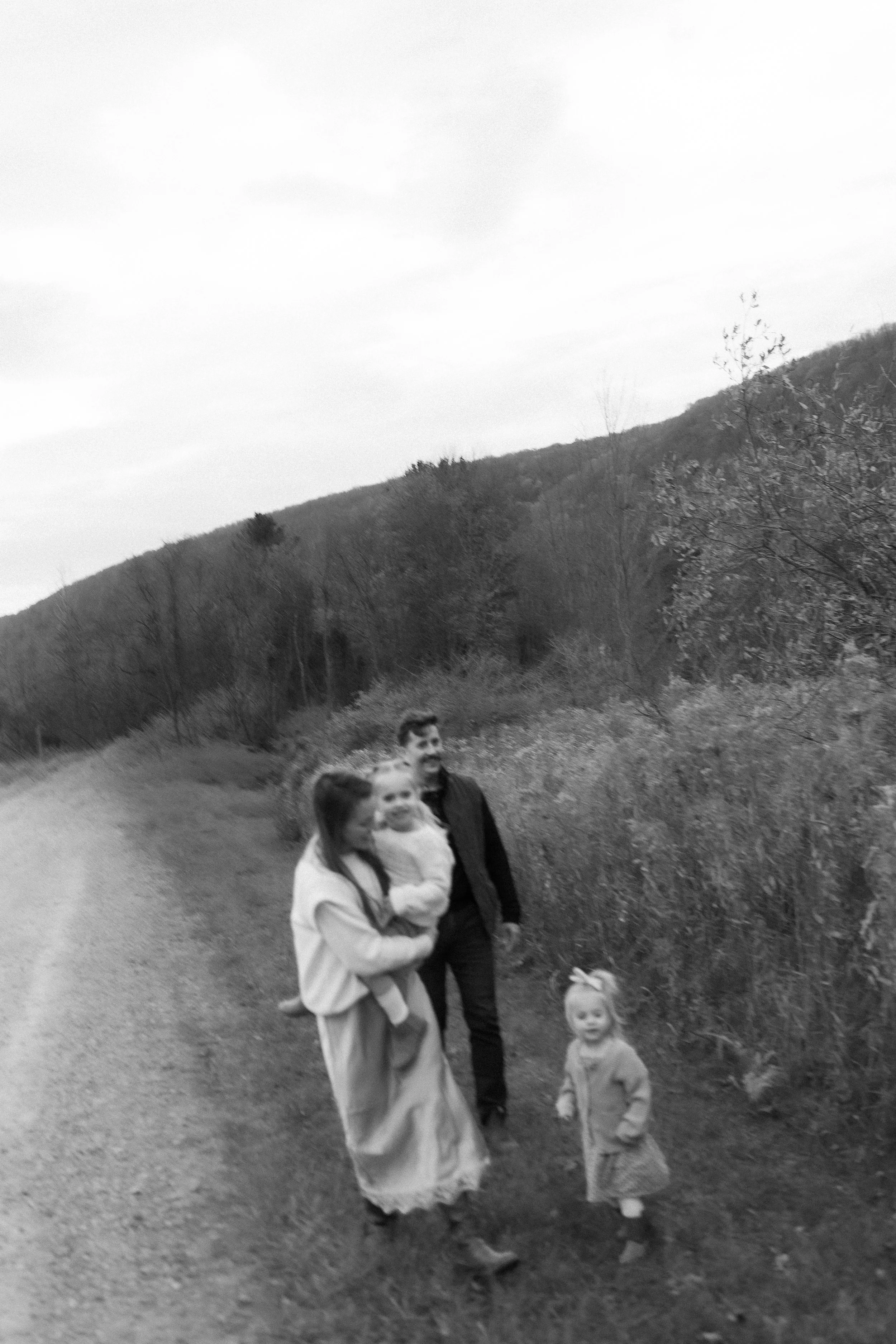 A family walking on a dirt path in a natural, outdoor setting with trees and hills in the background.