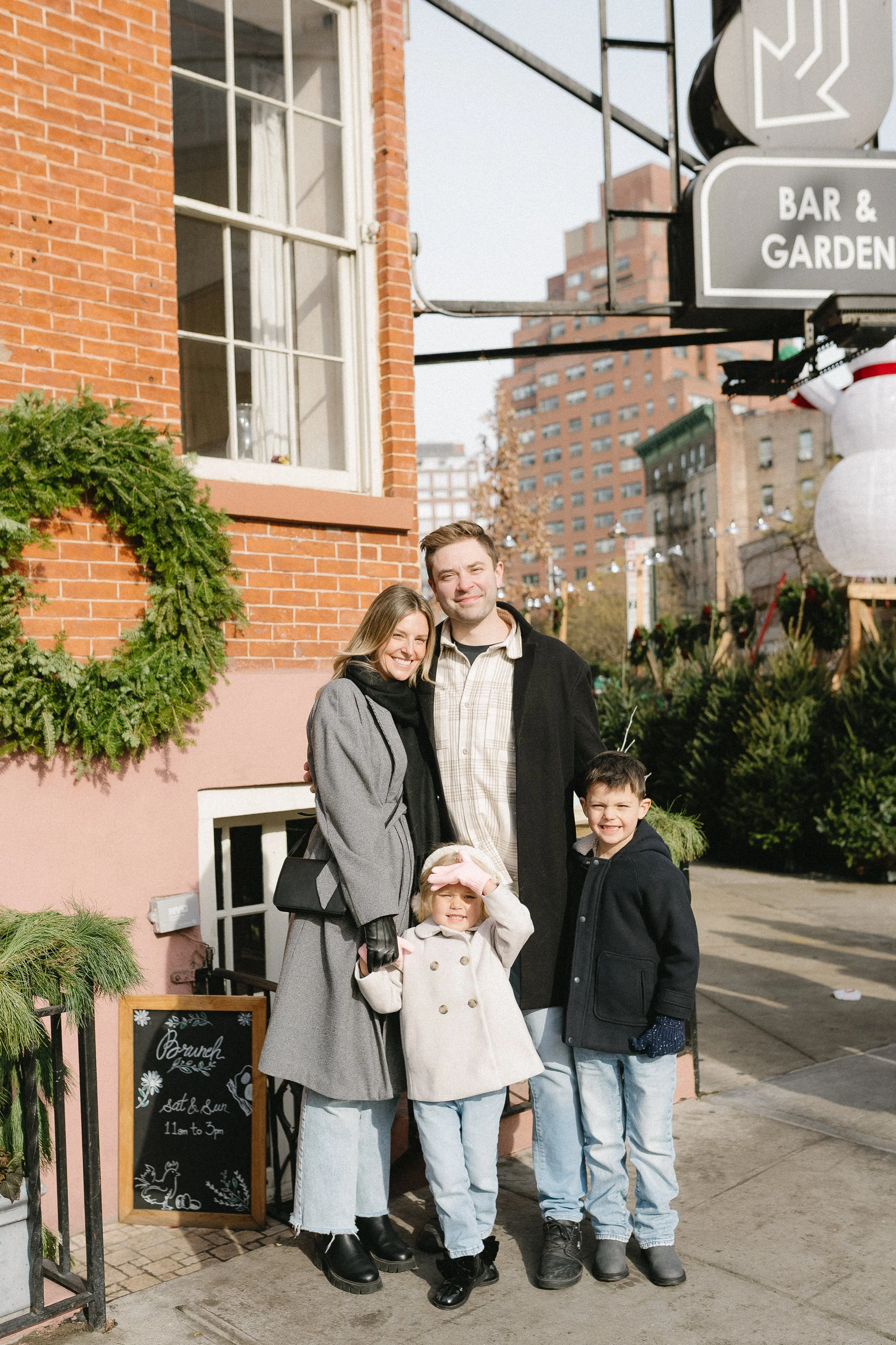 A family of four, including two children, stands outside a brick building decorated with greenery, smiling on a city sidewalk during daytime.