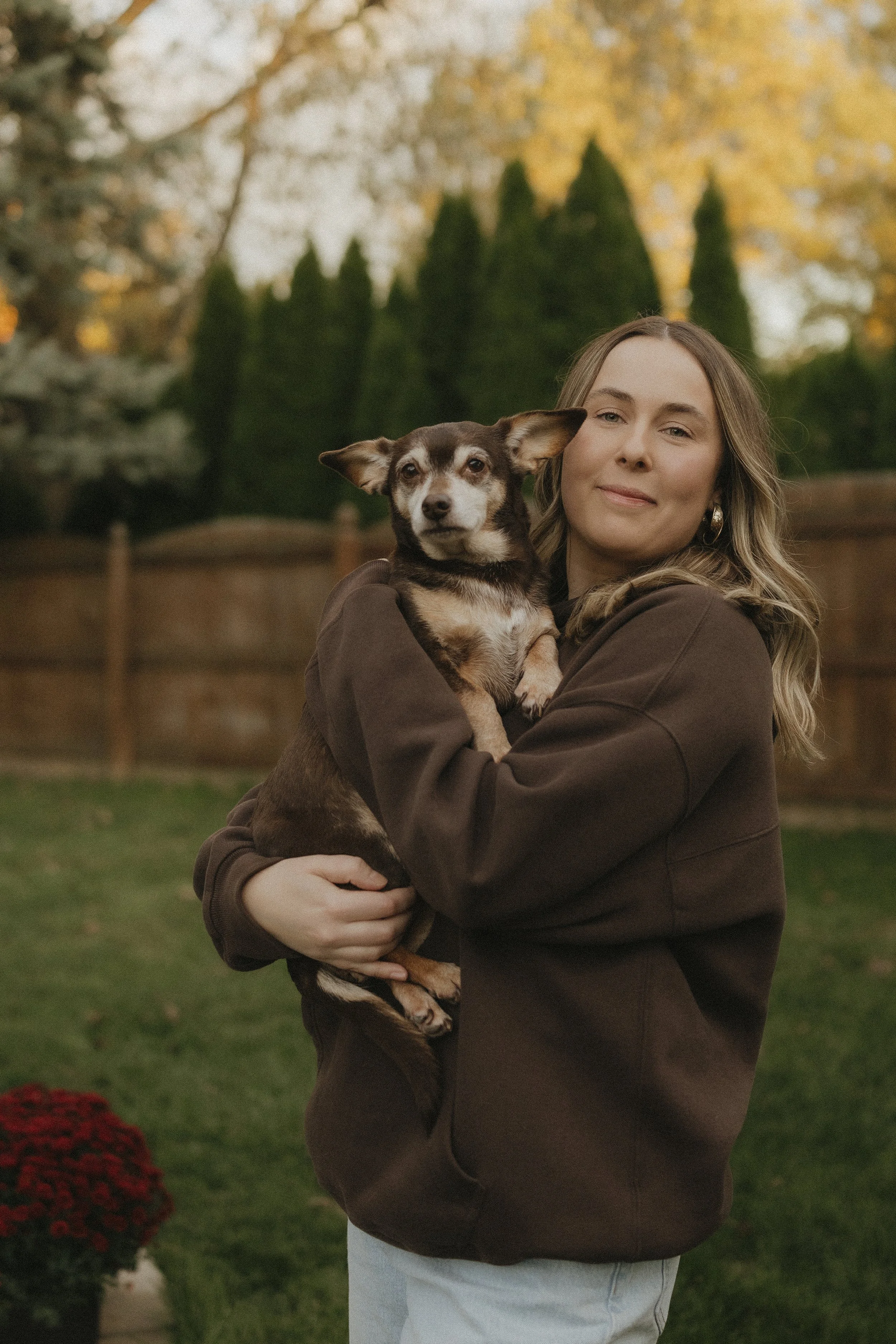 A young woman in a brown hoodie holding a small brown and black dog outdoors during fall, with trees and a wooden fence in the background.