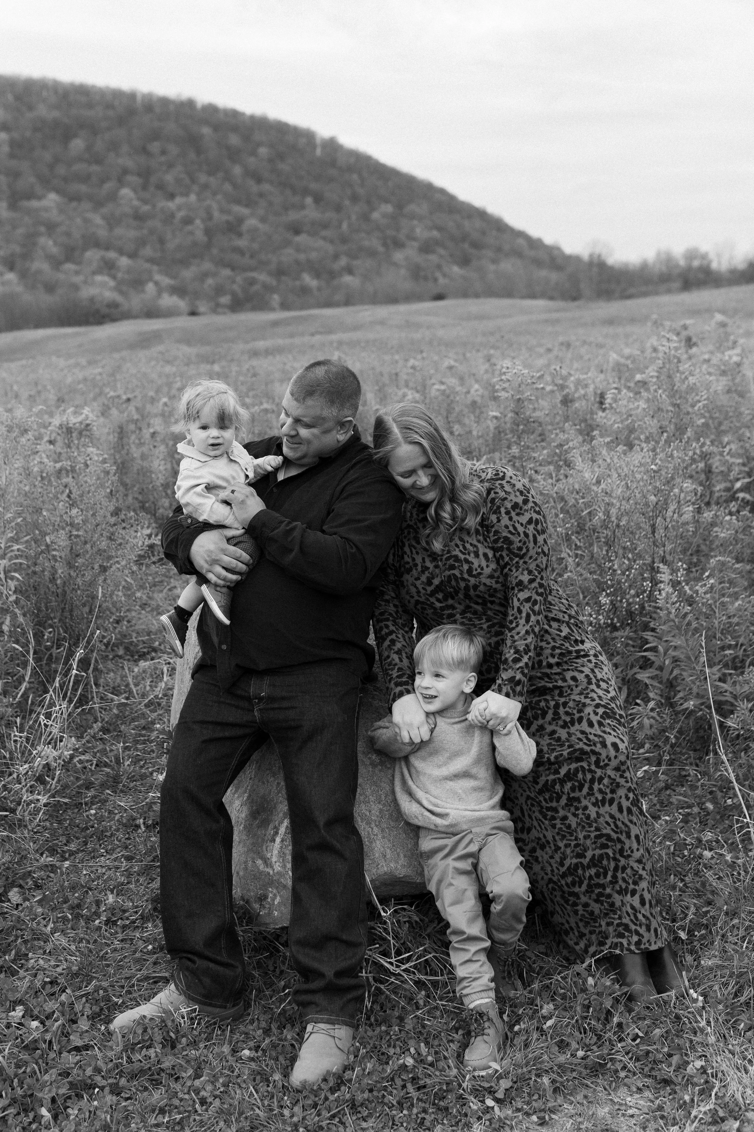 A family of four outdoors in a field with rolling hills in the background. The father holds a young girl, the mother is next to him, and a young boy is smiling and holding the mother's hand.