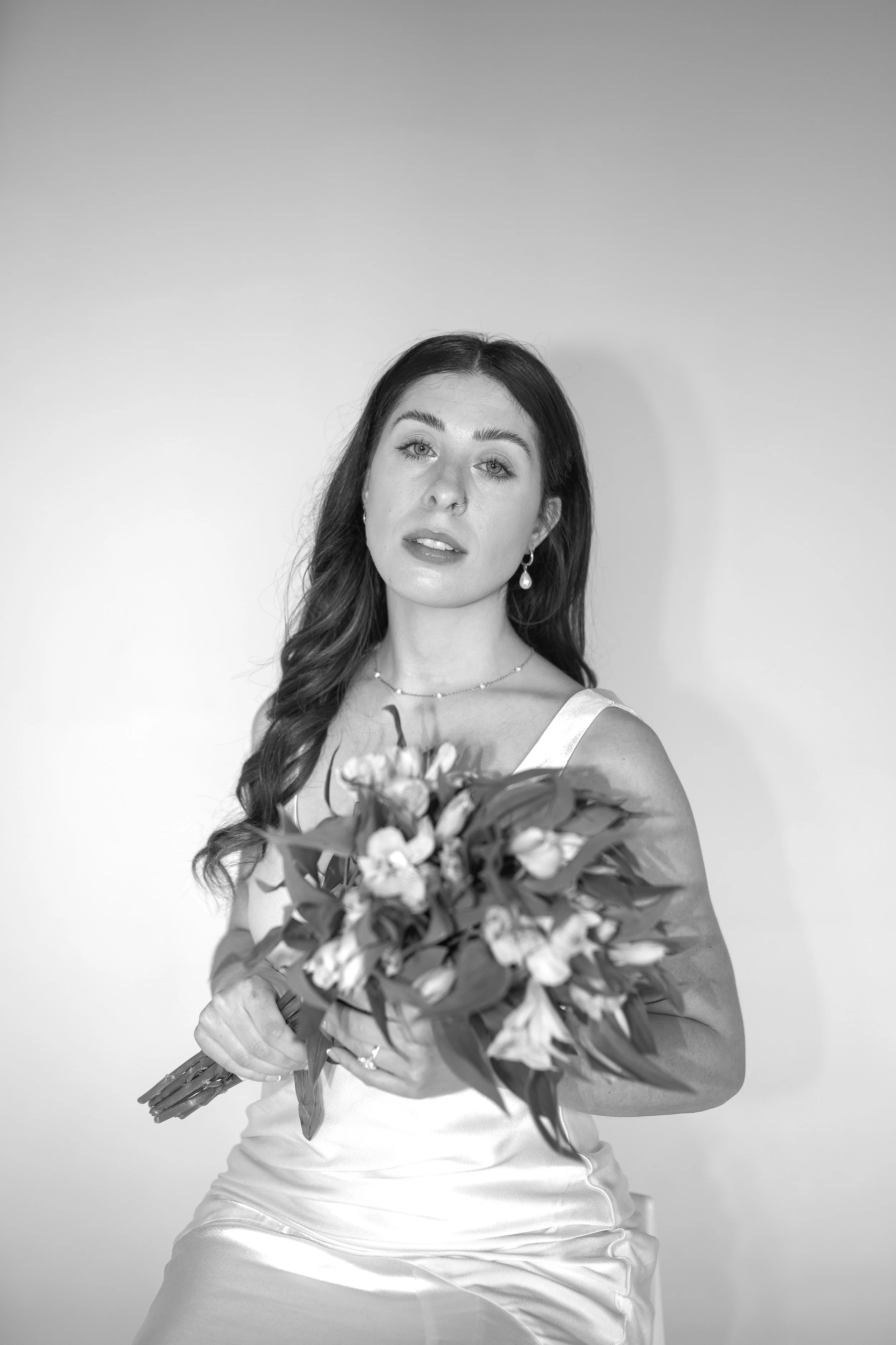 Black and white portrait of a woman with long wavy hair, wearing earrings, necklaces, and a satin dress, holding a bouquet of flowers, against a plain background.