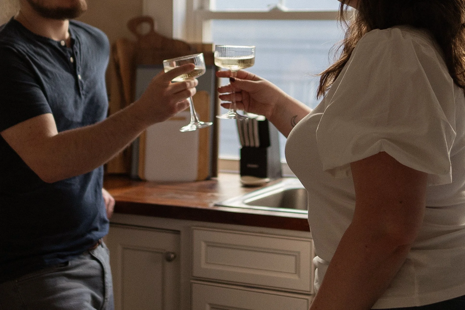 Two people clinking glasses of white wine in a kitchen.