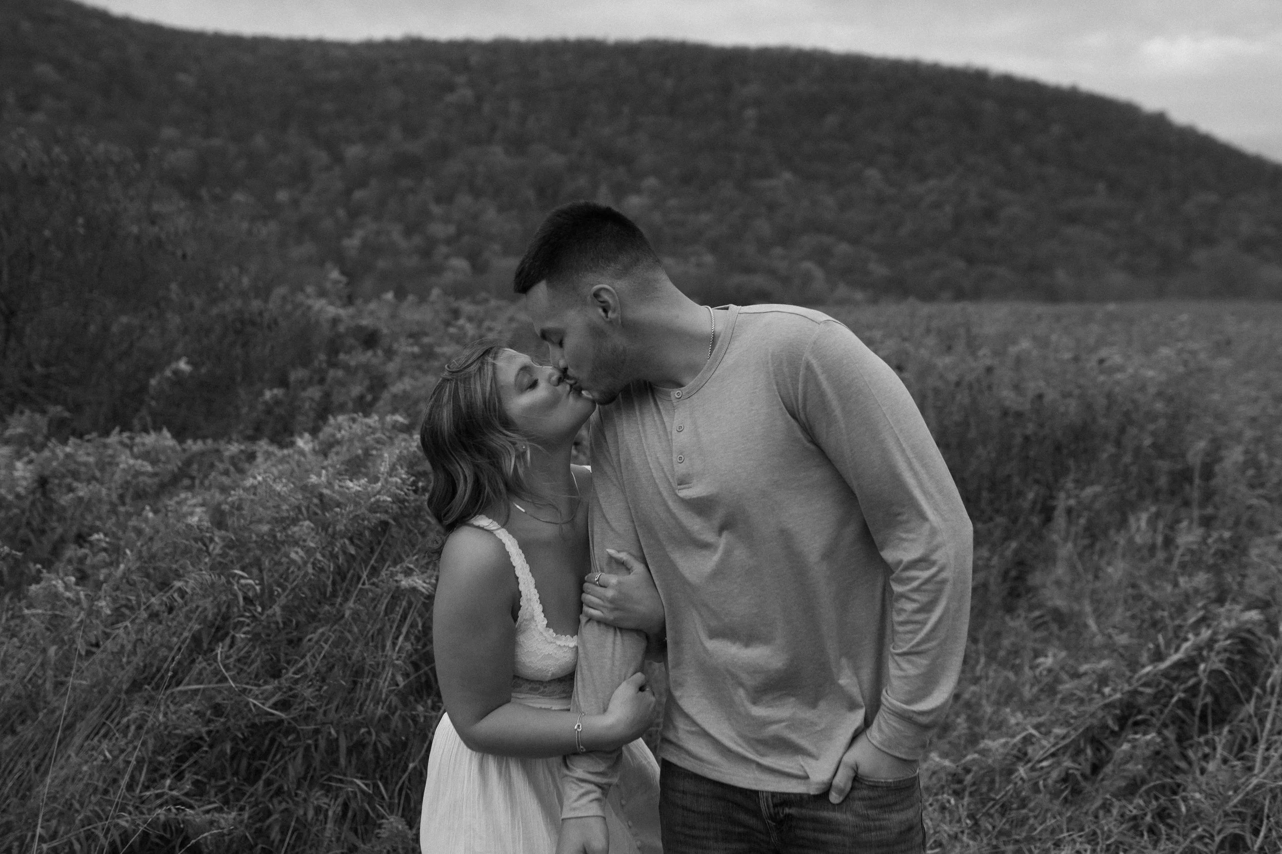 A young couple sharing a kiss outdoors in a field with hills in the background.