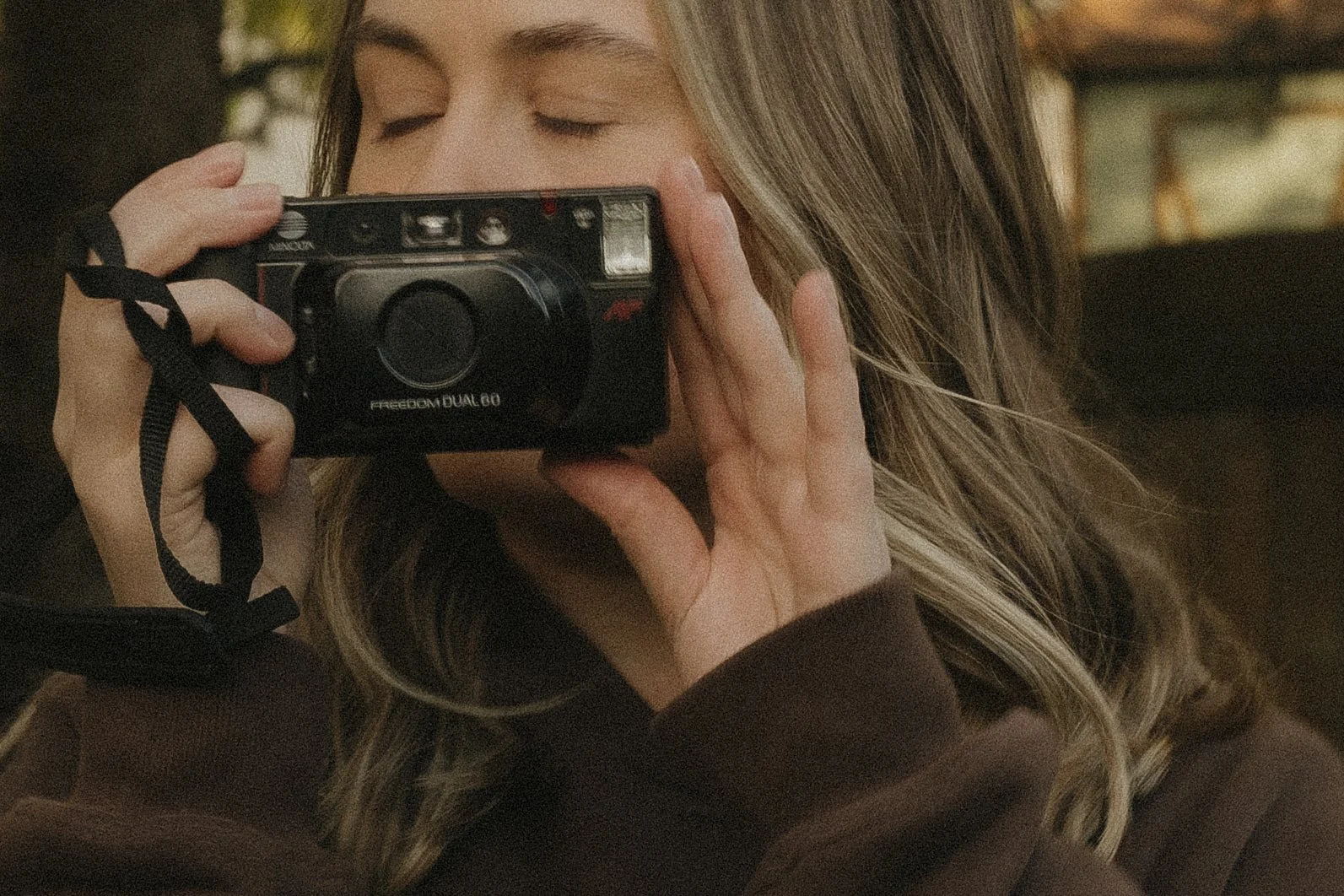 A woman with long blonde hair is holding a black camera up to her face with her eyes closed, taking a photograph. She is wearing a brown long-sleeve top.
