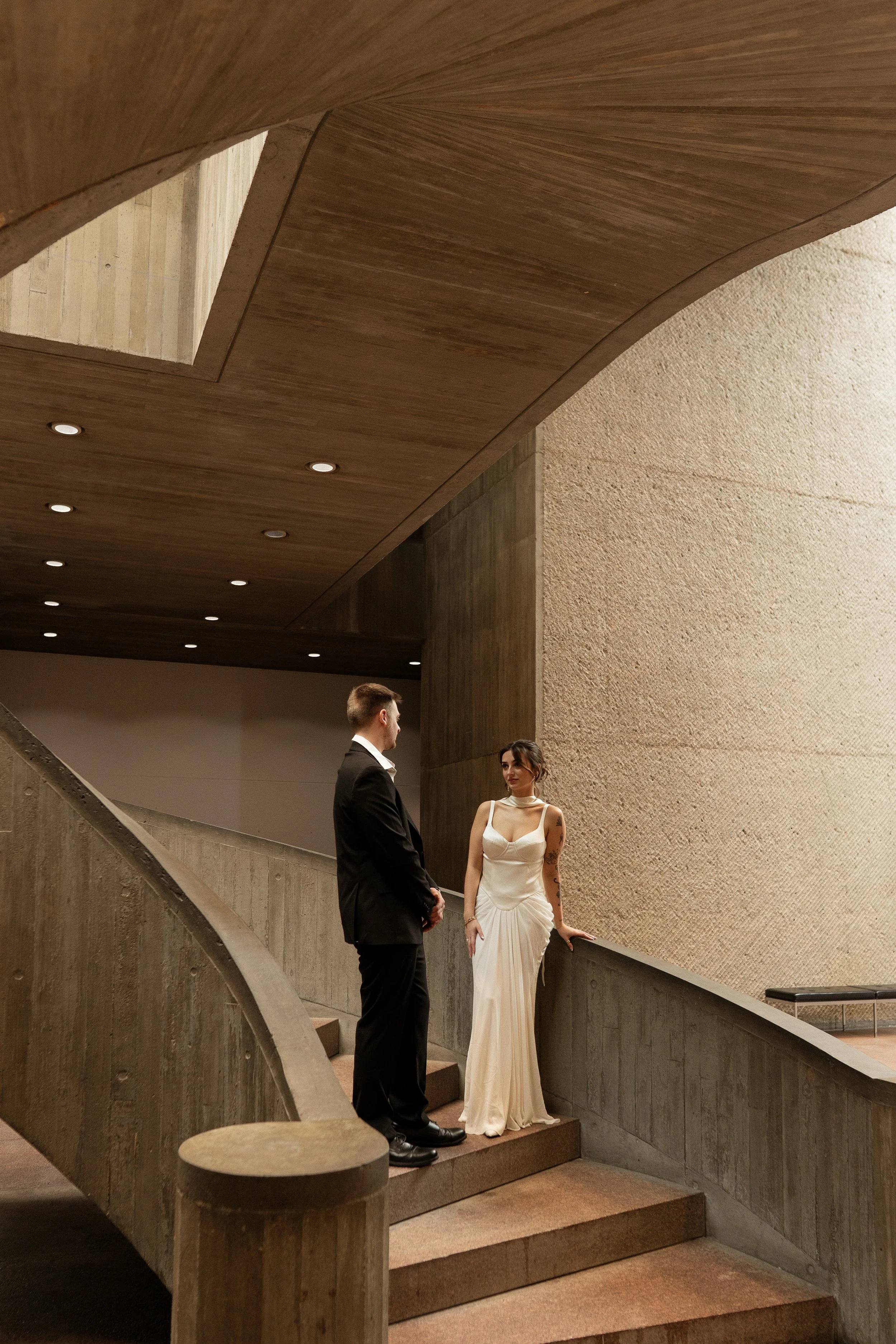A man and woman in formal attire standing on a staircase indoors, engaging in conversation. The woman is wearing a white gown, and the man is in a black suit. The setting has modern architectural elements with a textured wall and wooden ceiling.