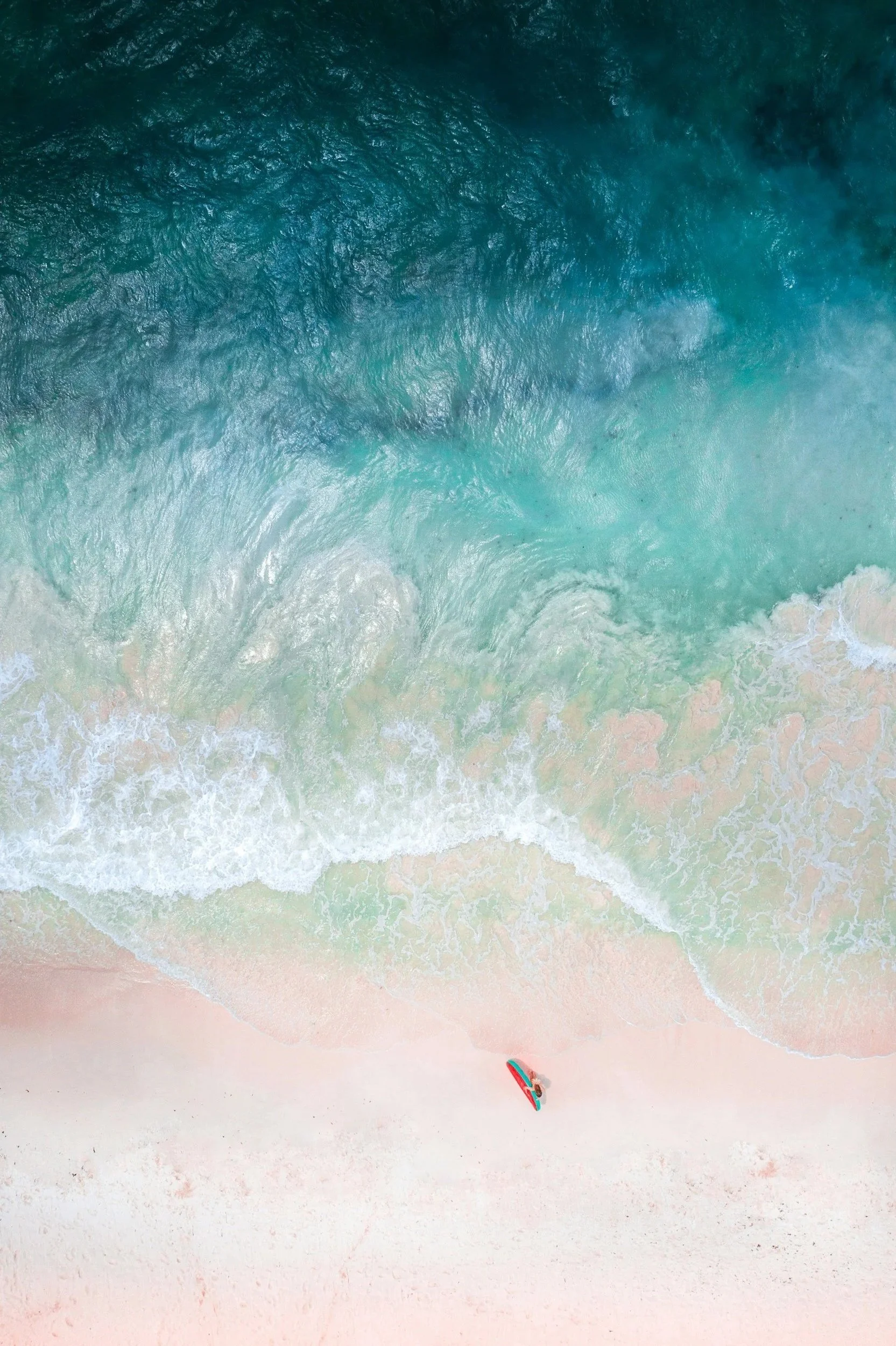 An aerial view of a beach with waves crashing on the shore, and a person standing on the sand with a surfboard.