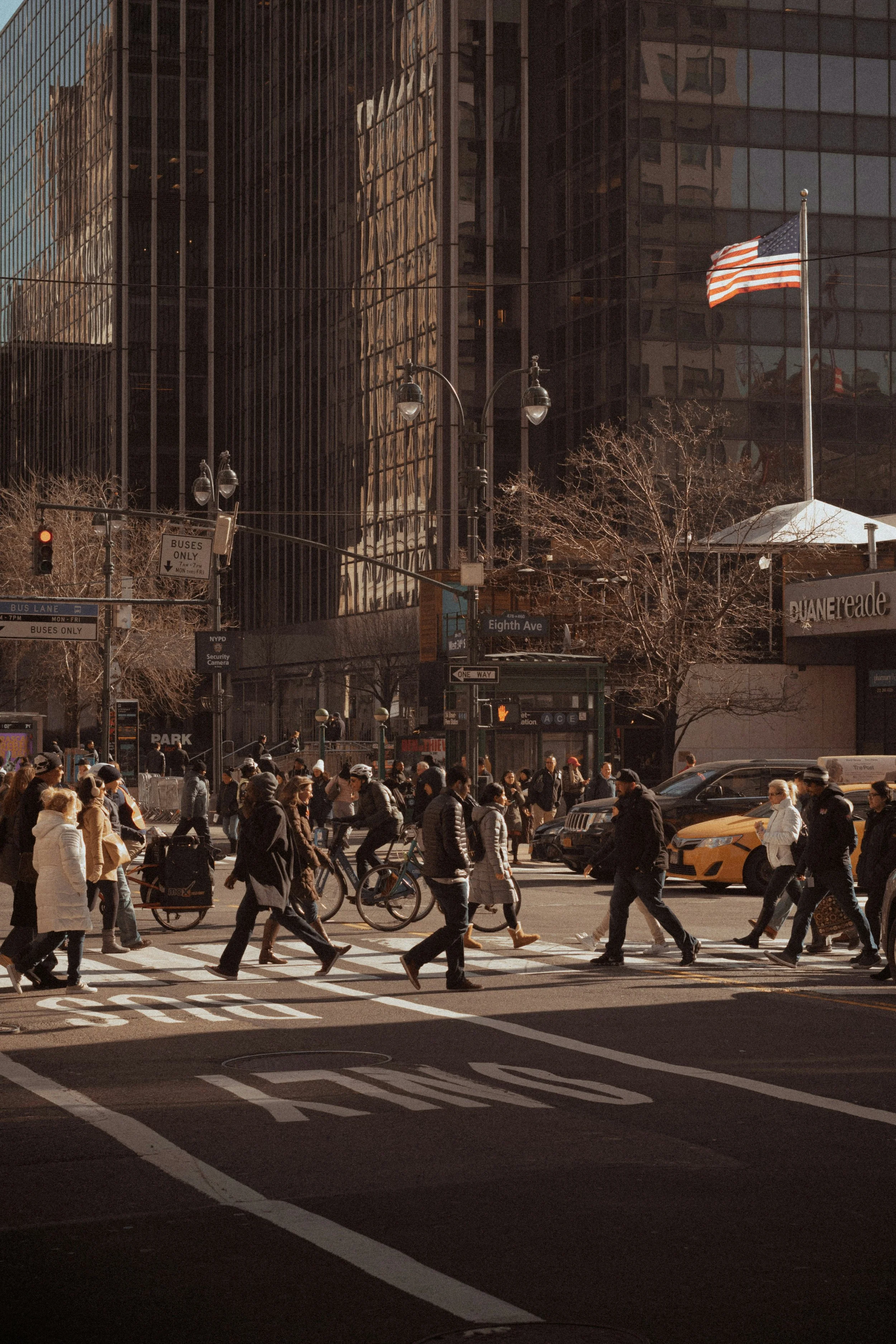 Crowded city street with pedestrians crossing at a crosswalk, tall office buildings, an American flag, and traffic signals in the background.