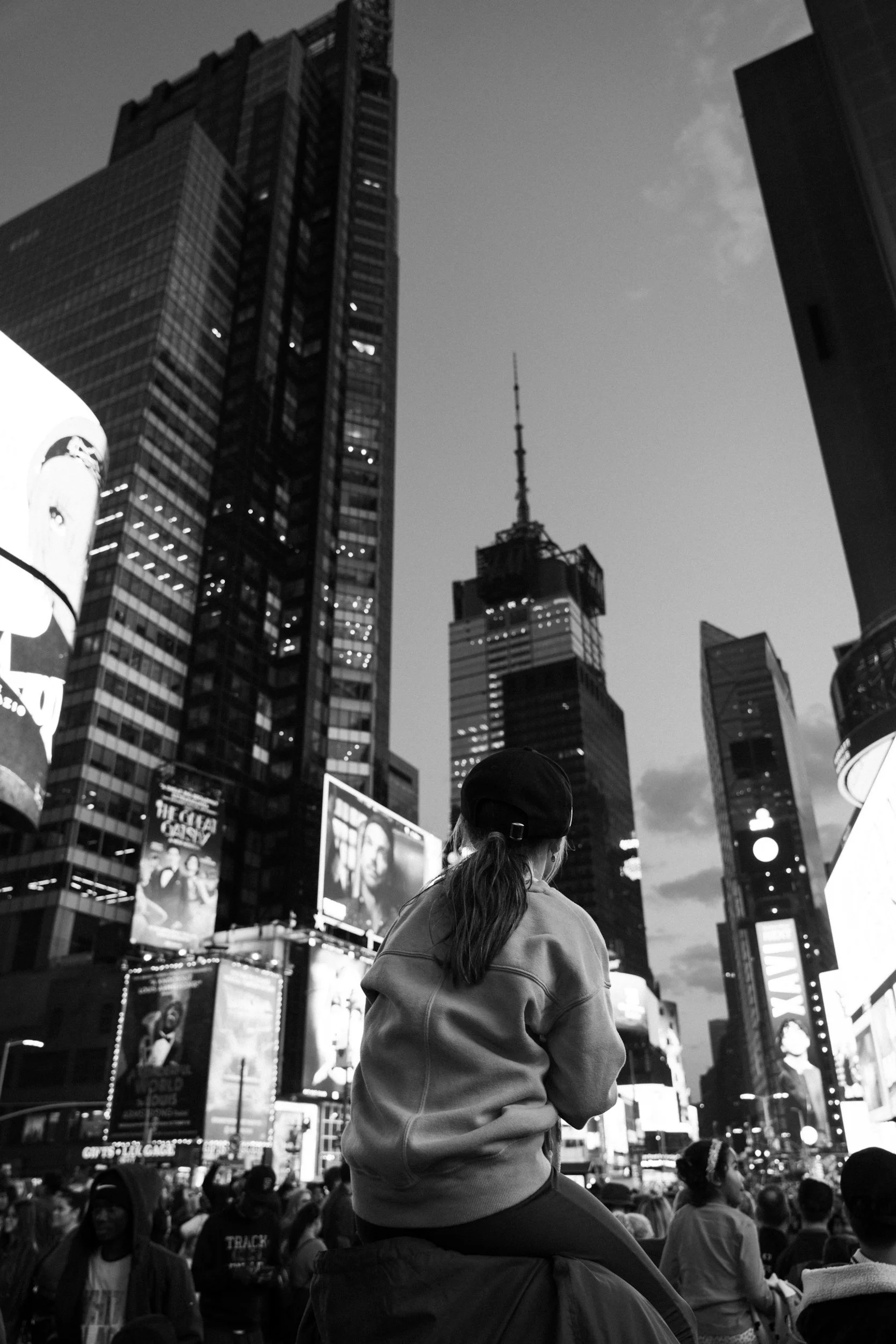 A woman with a ponytail wearing a beanie and oversized sweatshirt sitting on someone's shoulders in Times Square, New York City, surrounded by illuminated billboards and skyscrapers, including the One Times Square building with a spire.