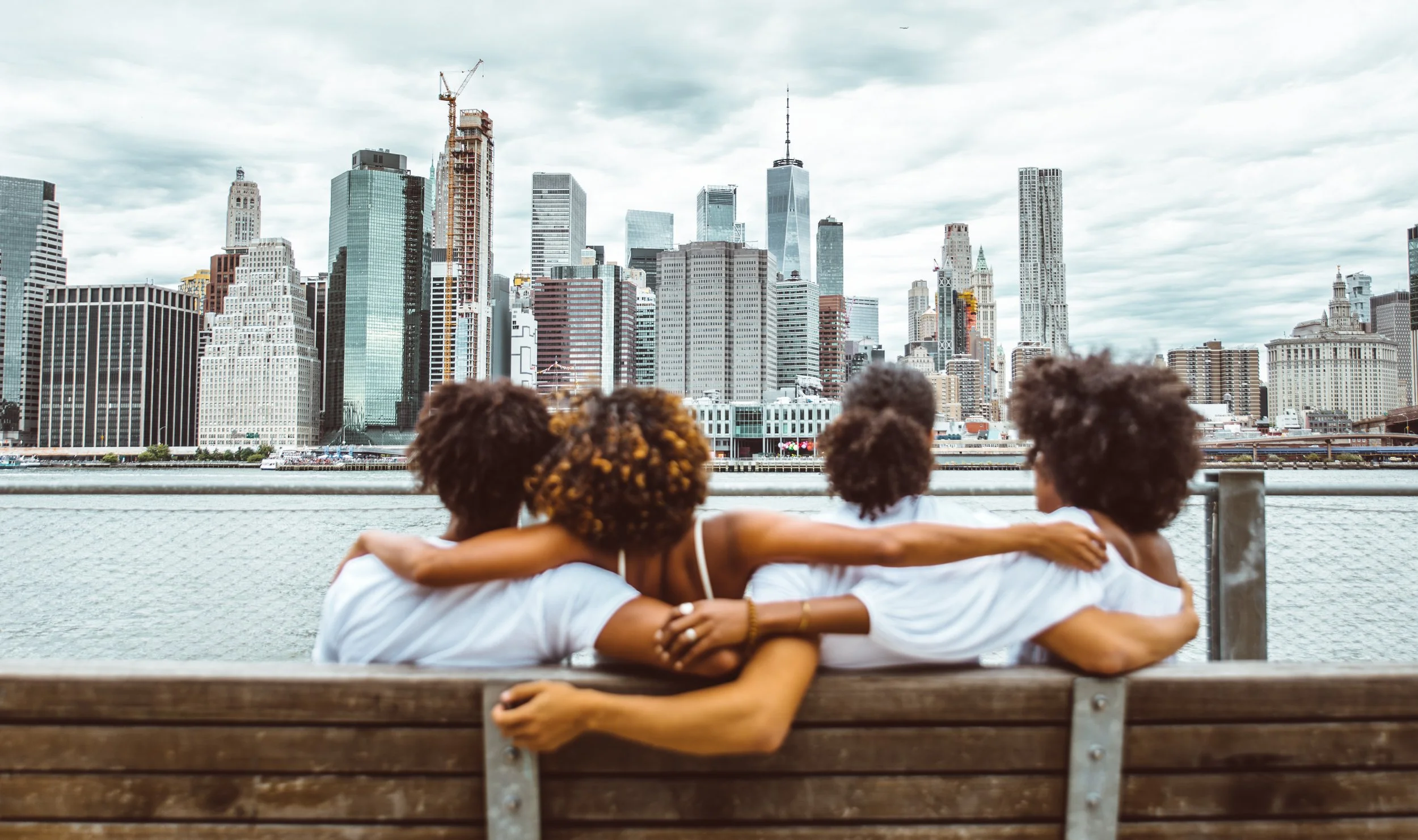 Four friends with curly hair sitting on a bench with their arms around each other, overlooking a city skyline with tall buildings and skyscrapers.