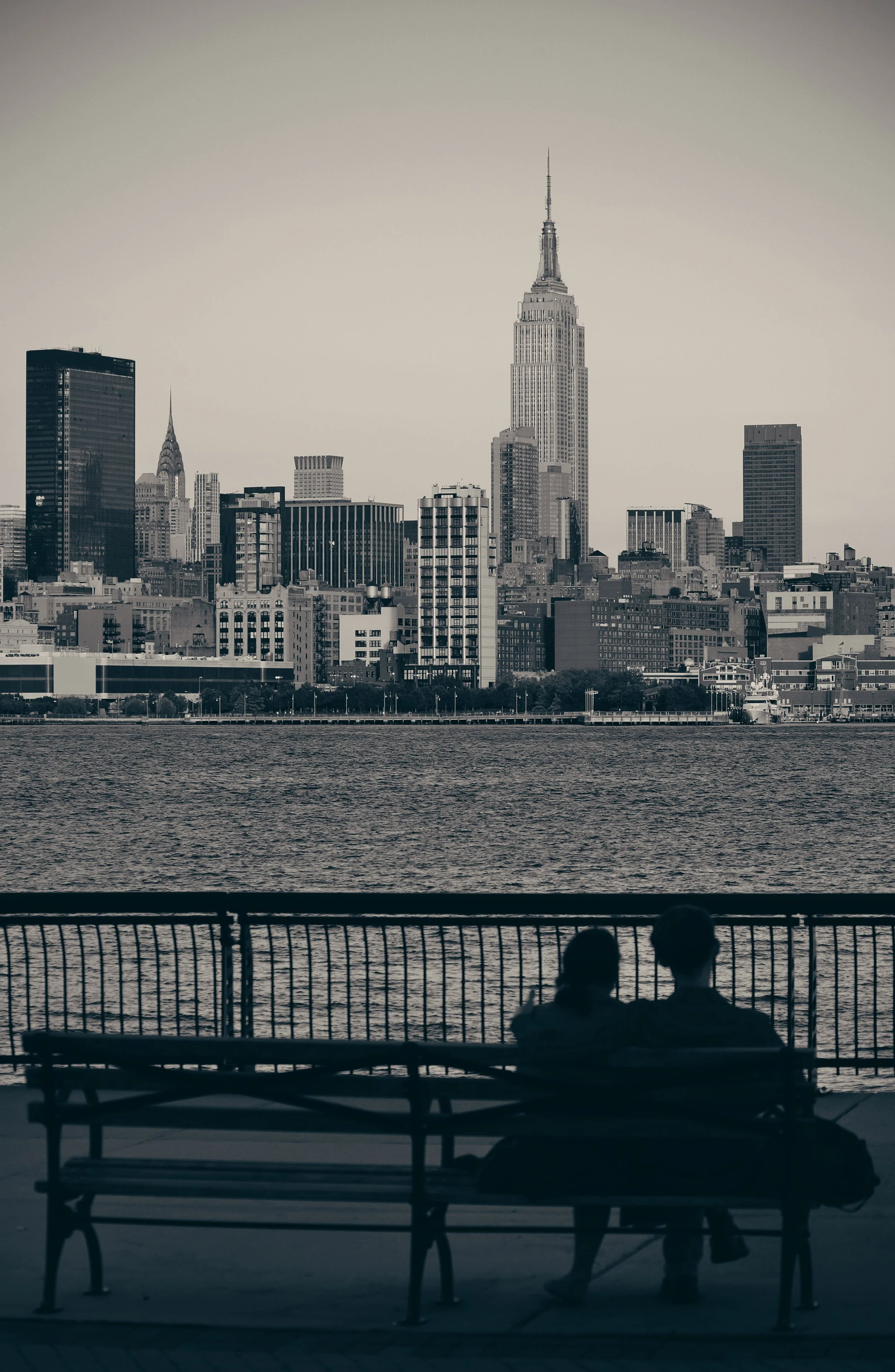 A black-and-white photo of two people sitting on benches by the water, overlooking the Manhattan skyline with the Empire State Building in the background.