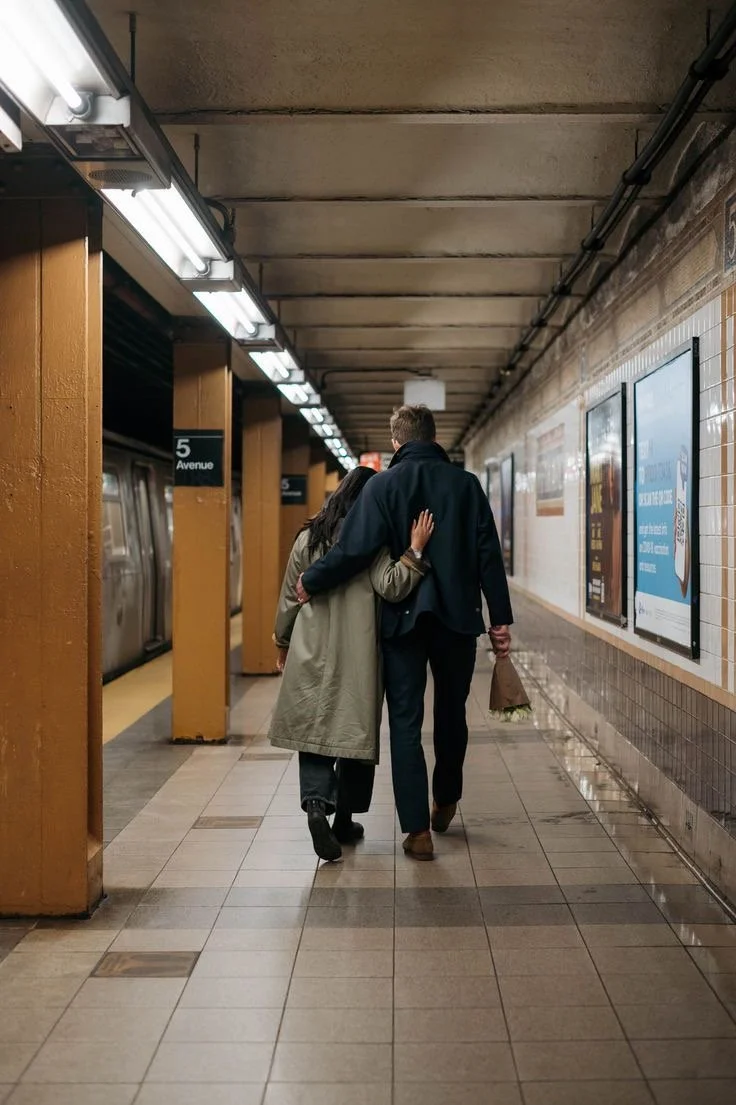 Two people walking together in a subway station, one with an arm around the other's shoulder, holding a shopping bag with the other hand.