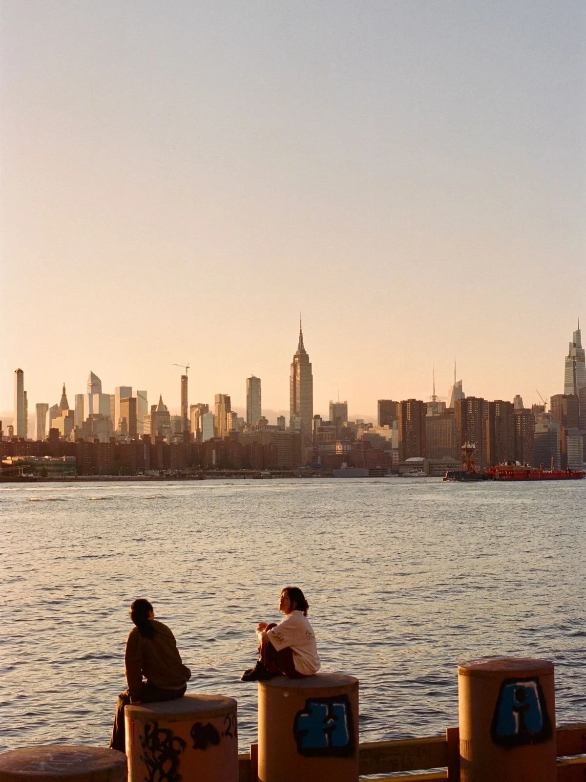 Two people sitting on concrete barriers by the water, with the New York City skyline, including the Empire State Building, in the background during sunset.