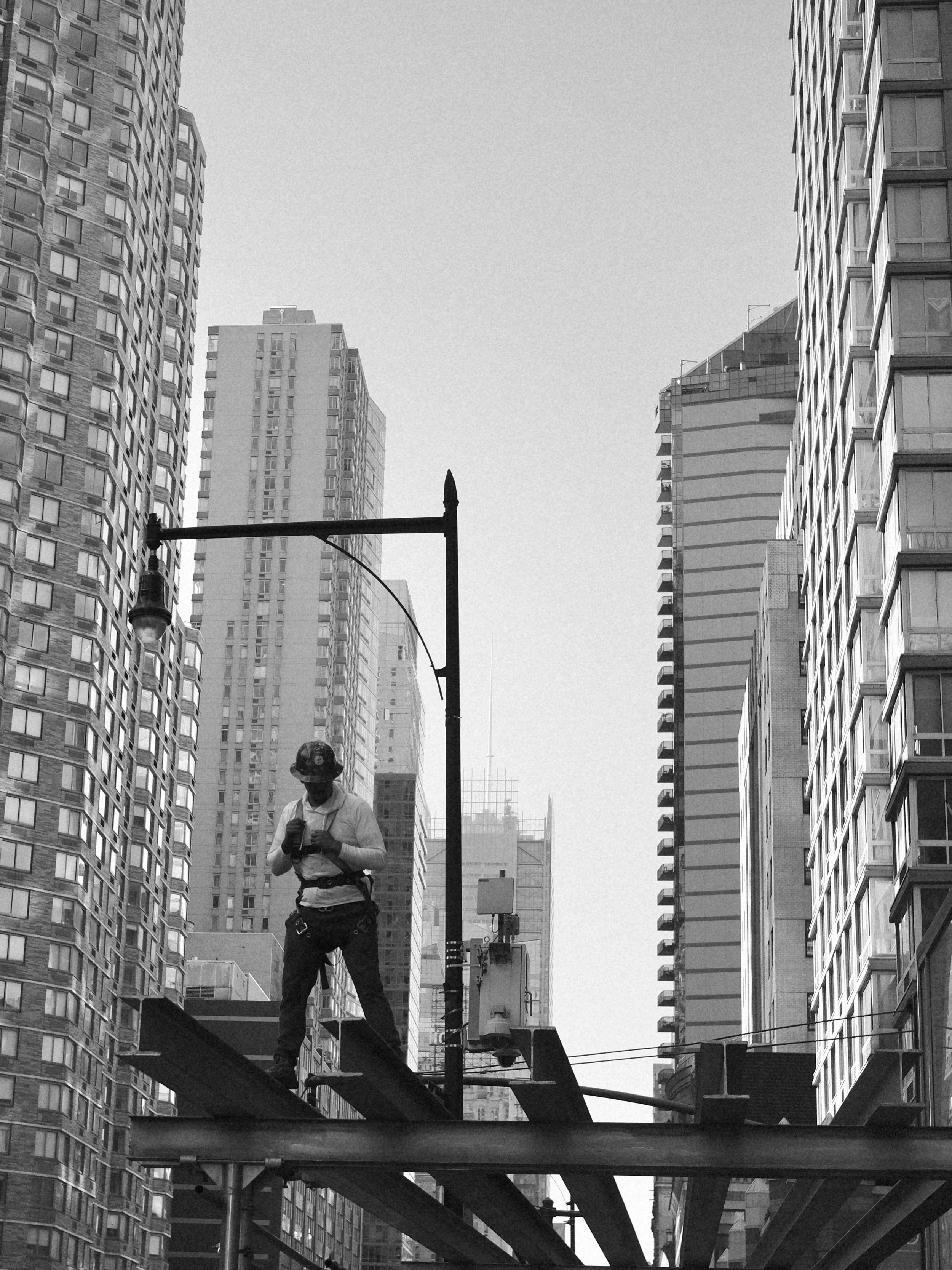 A worker wearing a hard hat and harness standing on steel beams in a city with tall buildings, using a device or phone.