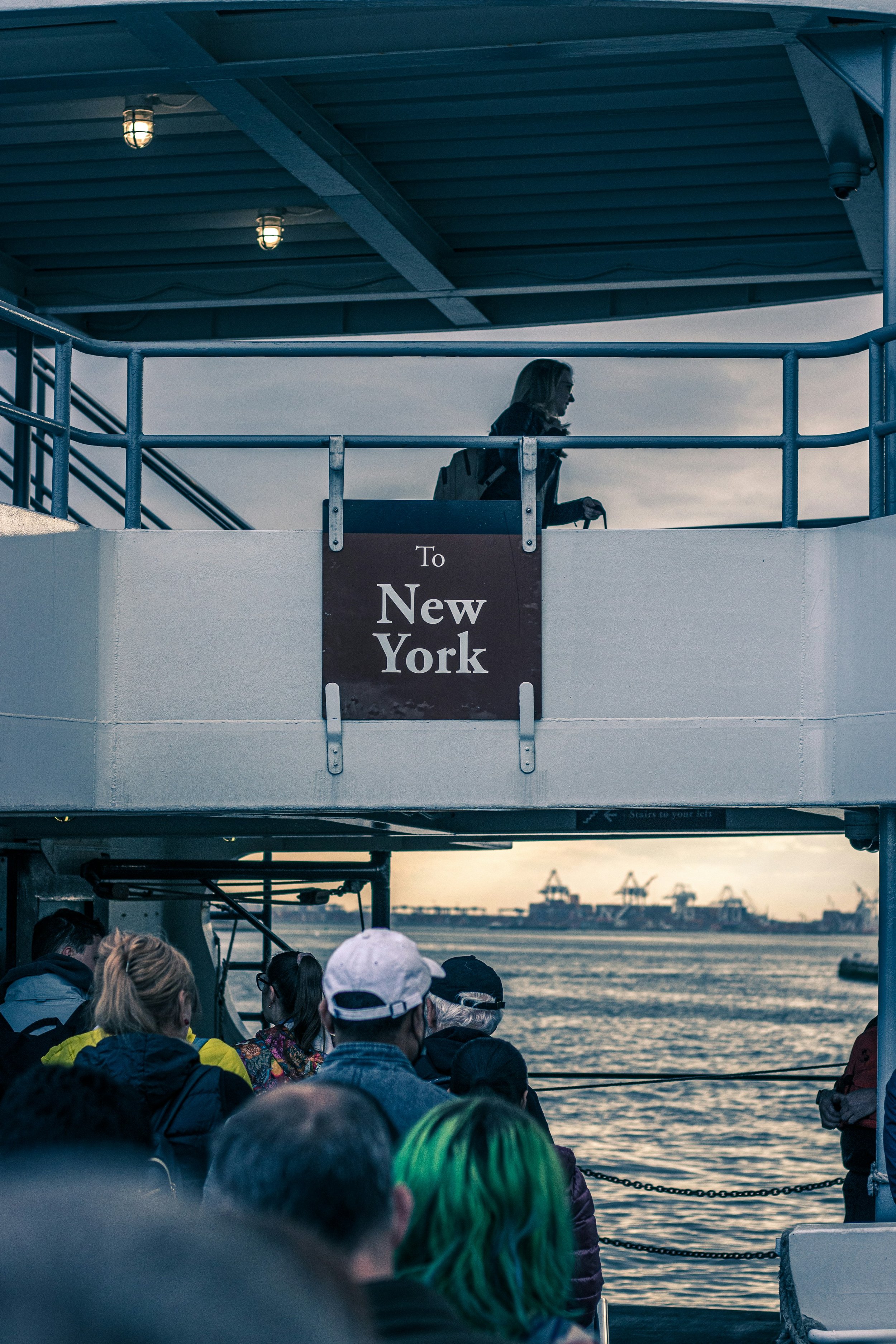 A woman walking on the upper deck of a ferry with a sign reading 'To New York' hanging on the railing, and a crowd of passengers on the lower deck looking at the water and the distant industrial port with cranes during sunset.