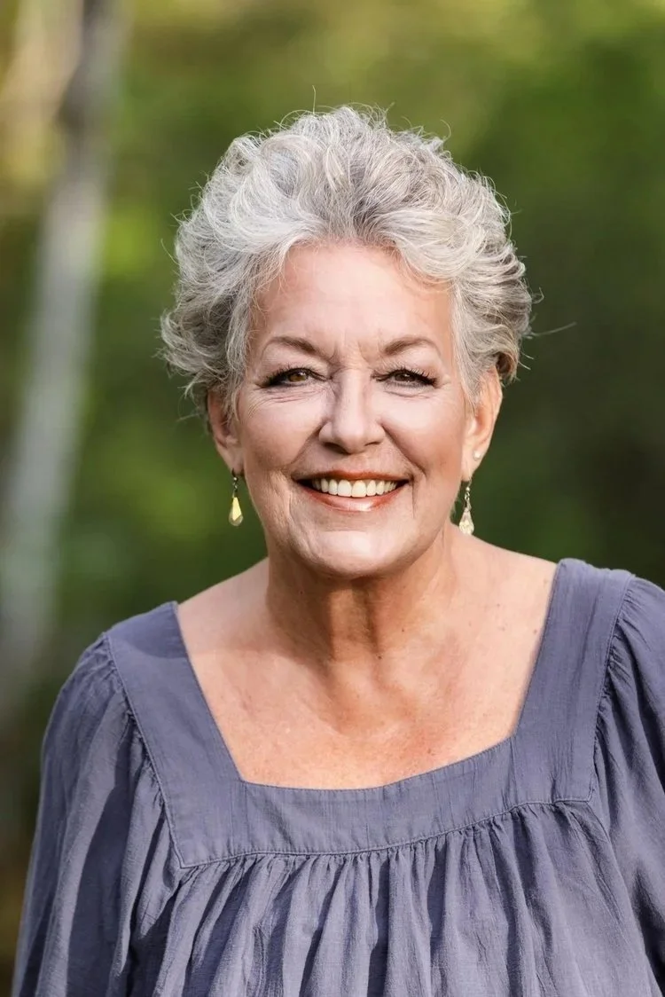 Close-up of an elderly woman with short gray hair smiling outdoors, wearing a purple blouse and earrings.