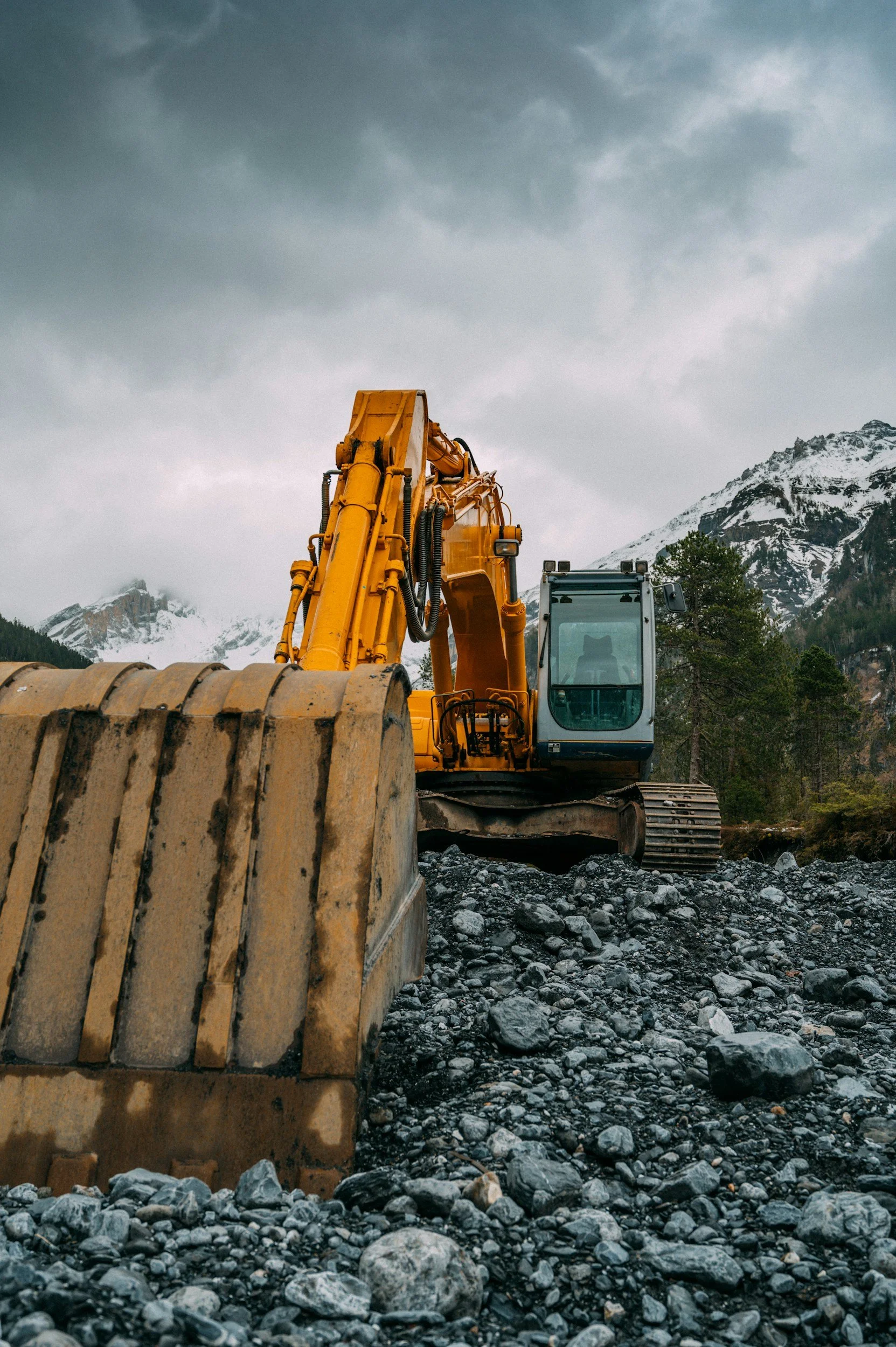 A yellow excavator on rocky terrain with snow-capped mountains and a cloudy sky in the background.