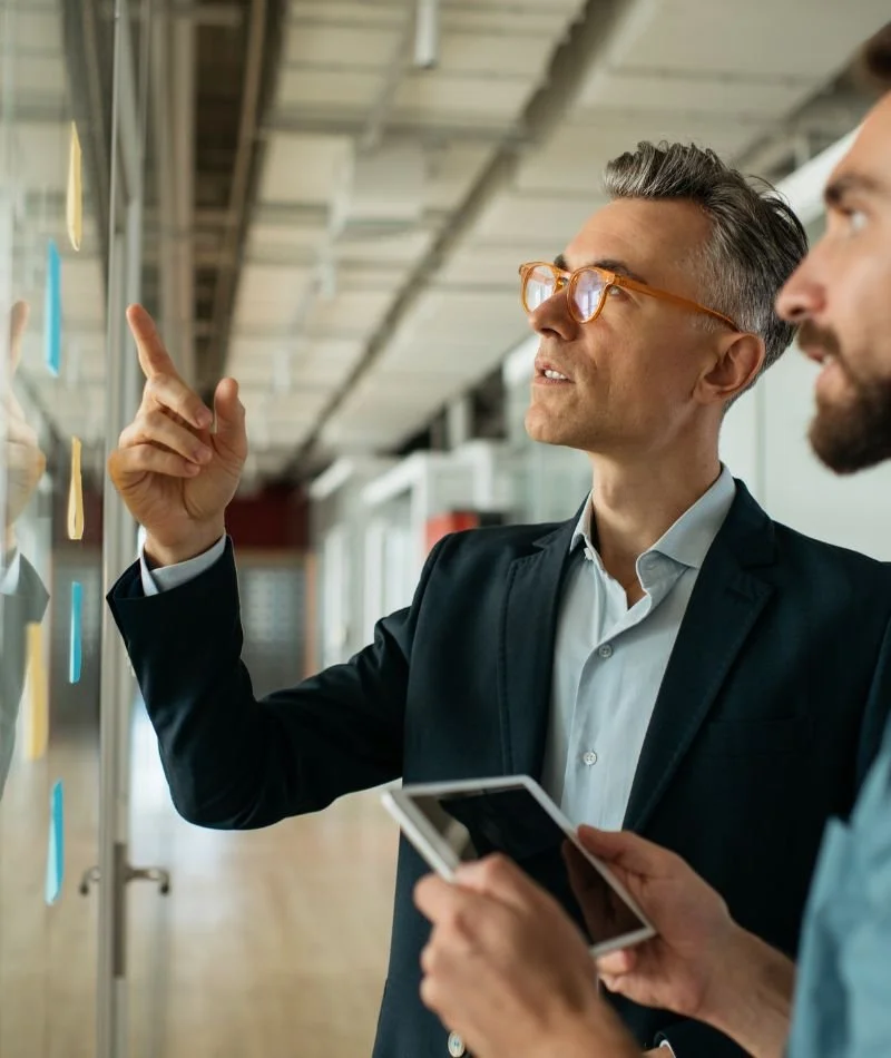 Two men in business attire having a discussion in a modern office or coworking space, with sticky notes on the glass wall.
