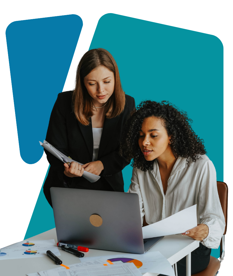 Two women working together at a desk with a laptop, papers, and markers.