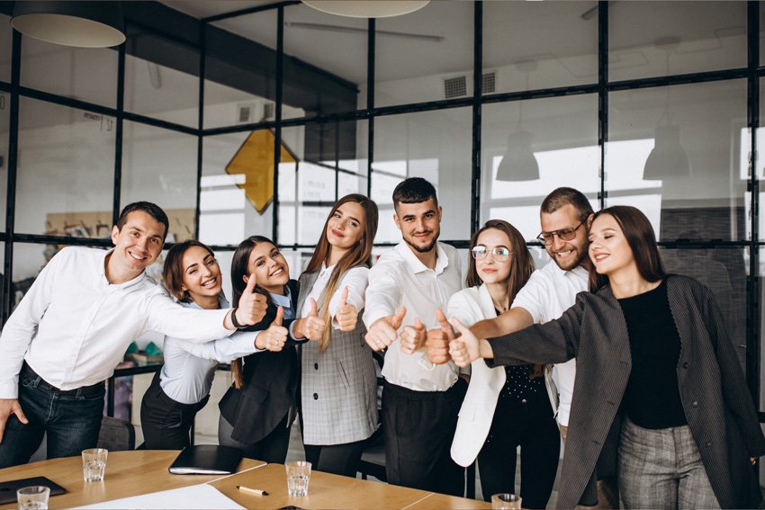 Group of eight diverse business professionals giving thumbs up in office conference room.