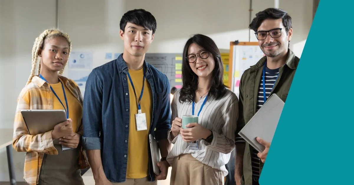 Group of four diverse young professionals standing together in an office, smiling, with charts and sticky notes on the wall behind them, holding notebooks and coffee cups.