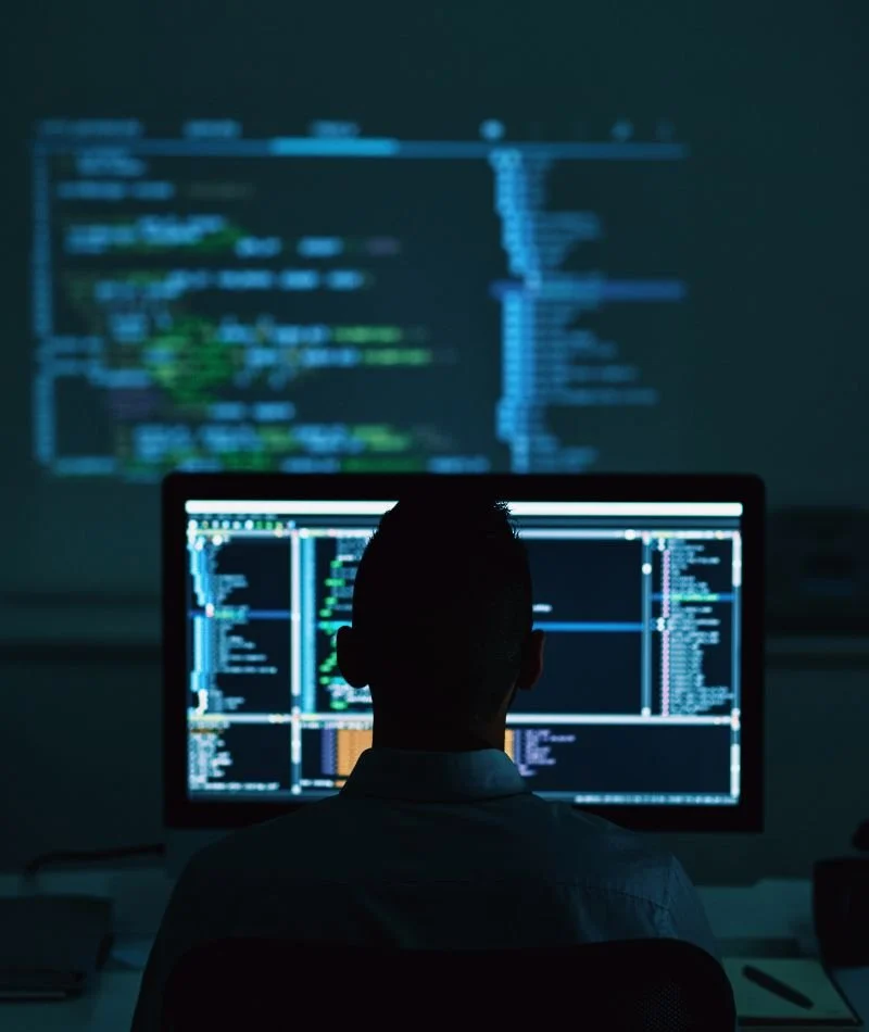 Person working on computer with code displayed on multiple screens, in a dark room with a projection of code on the wall behind.