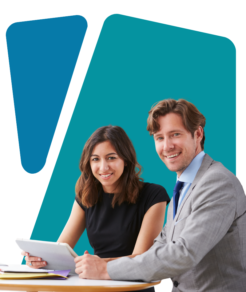 Two business professionals, a woman and a man, sitting at a table with documents and a tablet, smiling at the camera.