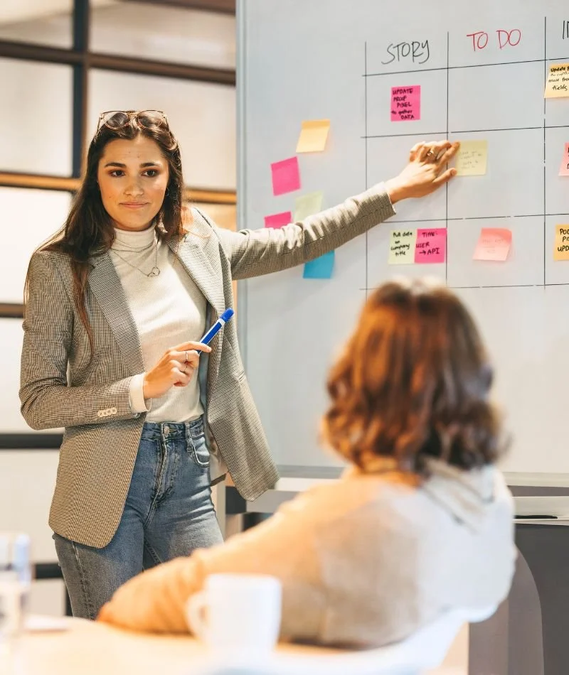 A woman in a blazer and jeans standing in front of a whiteboard with colorful sticky notes, pointing at the board during a meeting or presentation.