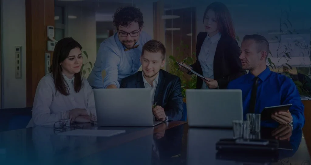Five professionals gathered around a conference table with laptops and tablets, collaborating in an office setting.