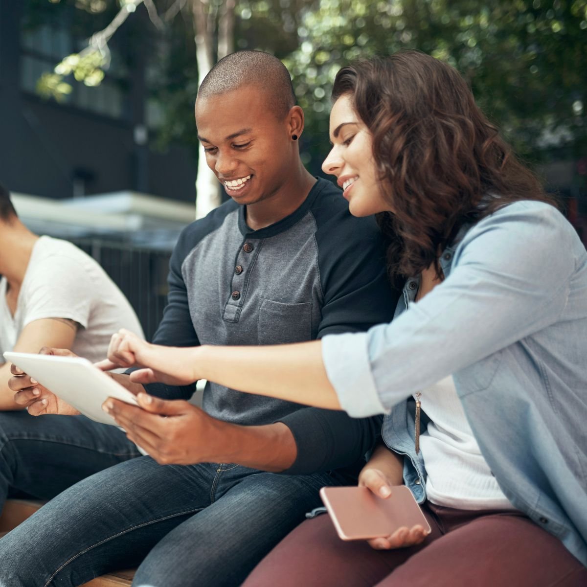 Two young adults, a man and a woman, sitting outdoors on a bench, looking at a tablet together and smiling, with a woman in the background using her phone.