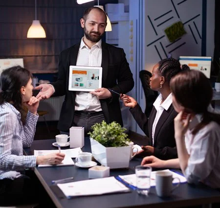 Business meeting with a man presenting a tablet screen to three women seated at a conference table in an office.