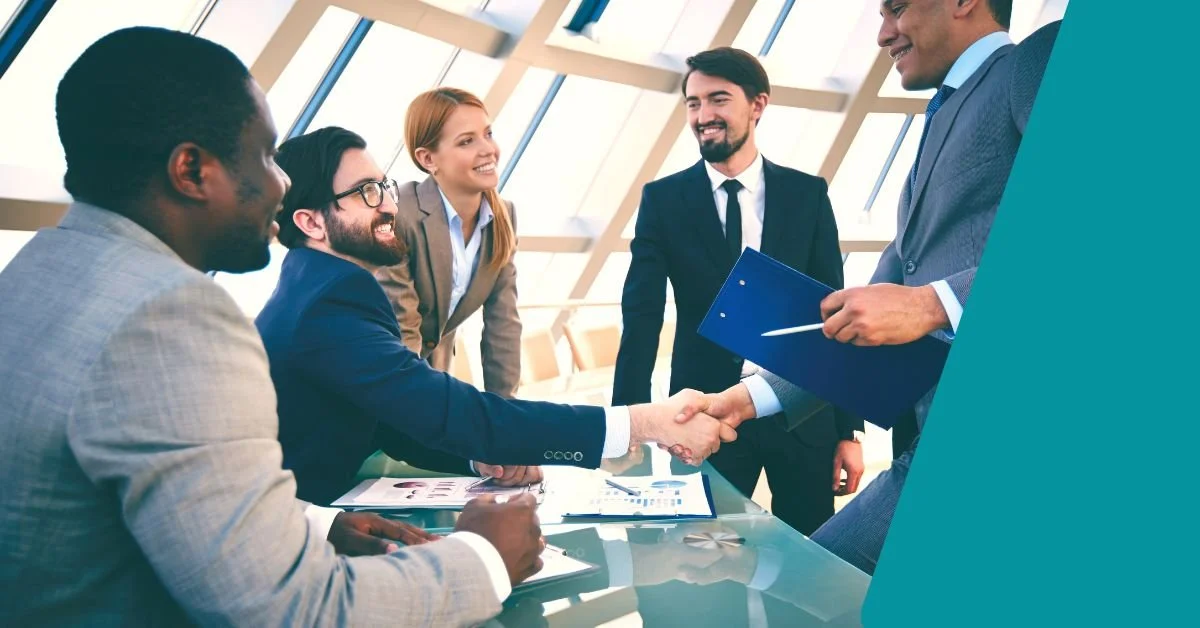 Business professionals in a meeting shaking hands, with laptops and documents on the table, in a modern office setting.