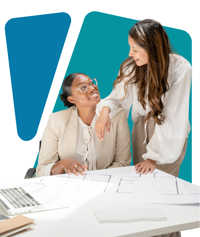 Two women having a friendly conversation at a table with architectural plans, one sitting and one leaning over smiling.