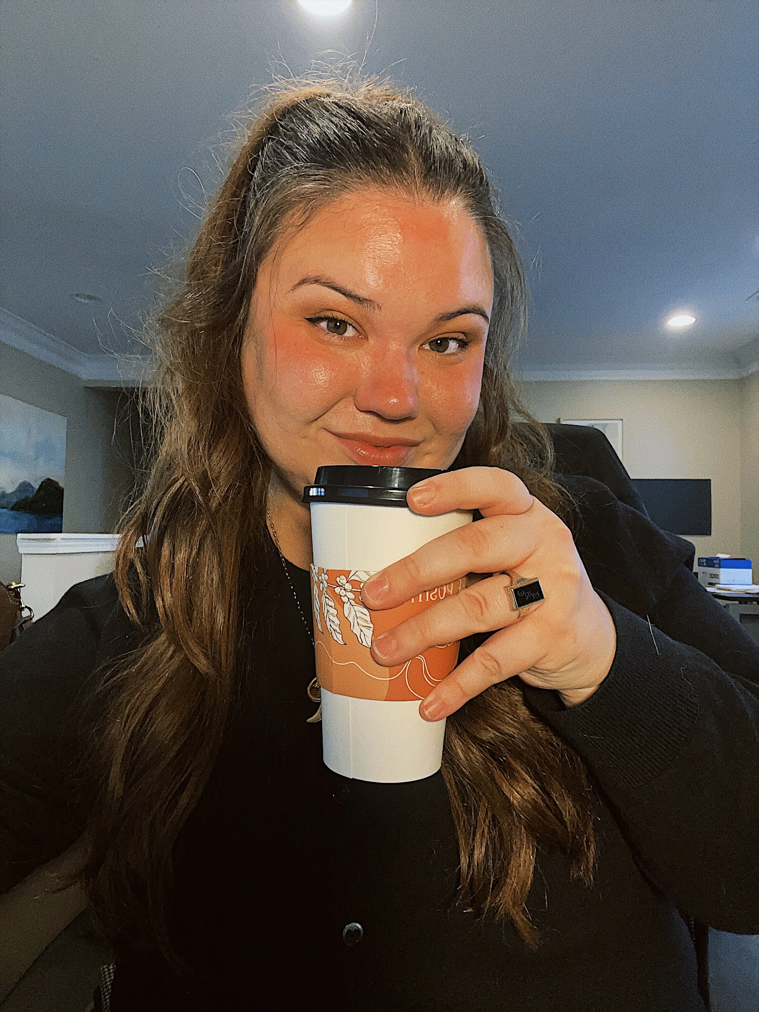 A woman with long brown hair and a black coat holding a travel mug, smiling at the camera indoors.