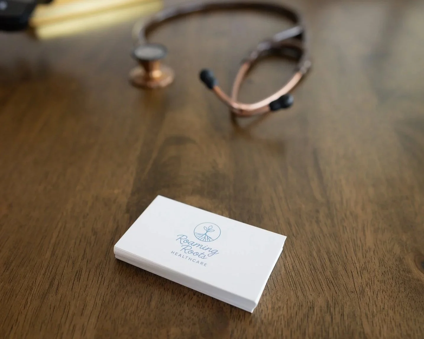 A stack of business cards with a logo of a plant and the text "Roaming Roots Healthcare," placed on a wooden table. In the background, a stethoscope is partly visible.