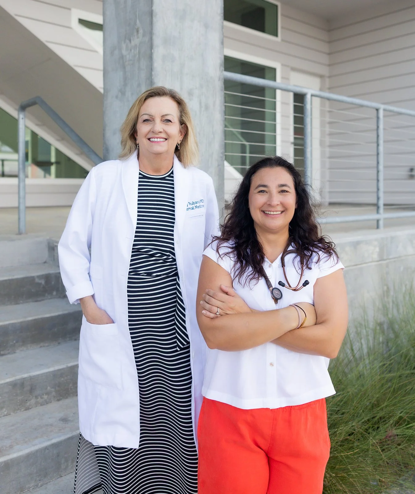 Two women, one wearing a white lab coat and the other with a stethoscope around her neck, stand outdoors in front of a modern building, smiling at the camera.