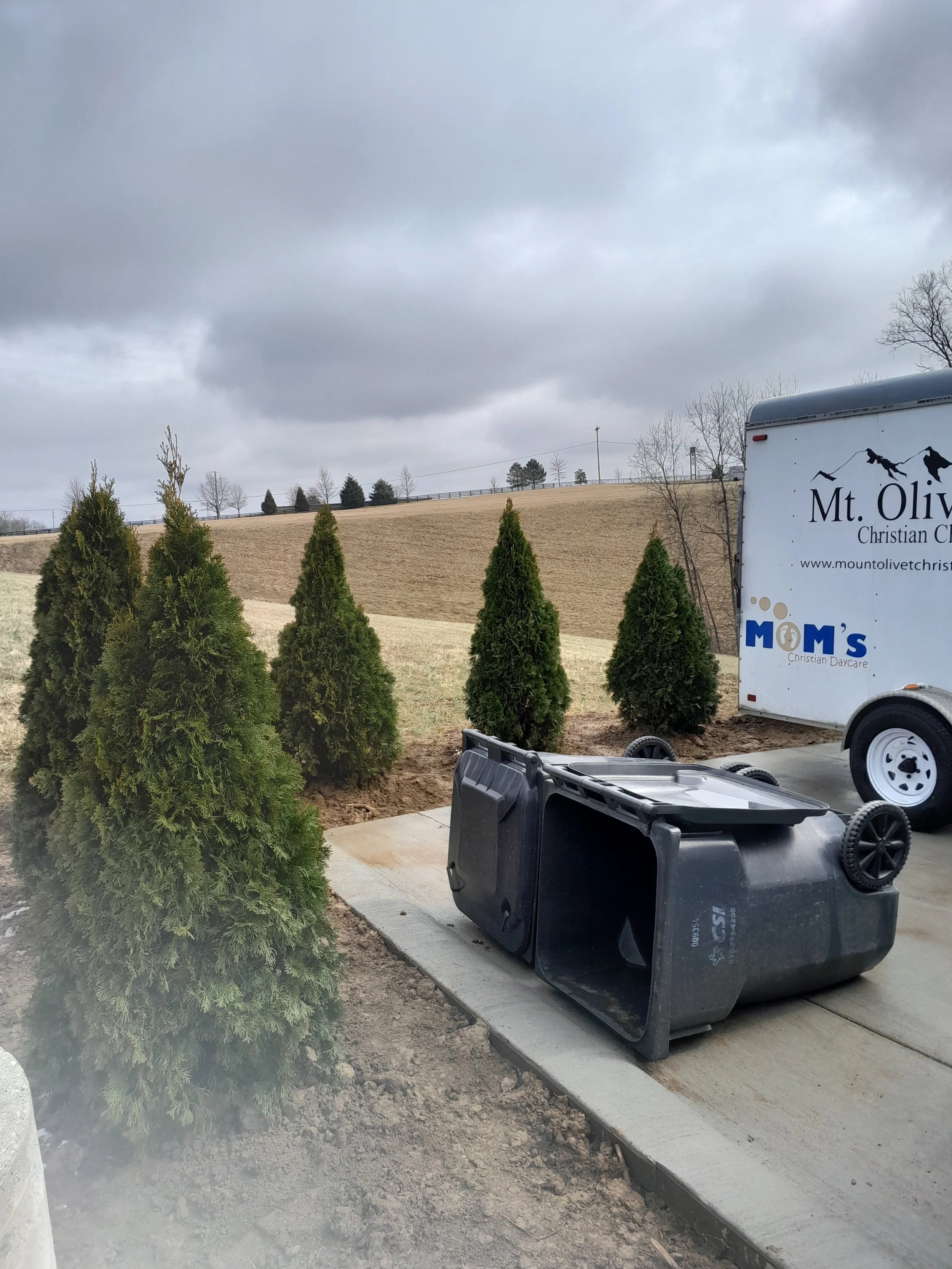 Upside-down trash bin on a sidewalk beside young evergreen trees and a trailer with a church logo at Mt. Olive Christian Church, with cloudy sky and open field in the background.