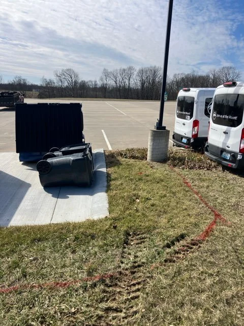 Empty parking lot with a few vans parked, a black trash bin, and a sidewalk on grass with tire tracks and red painted lines.
