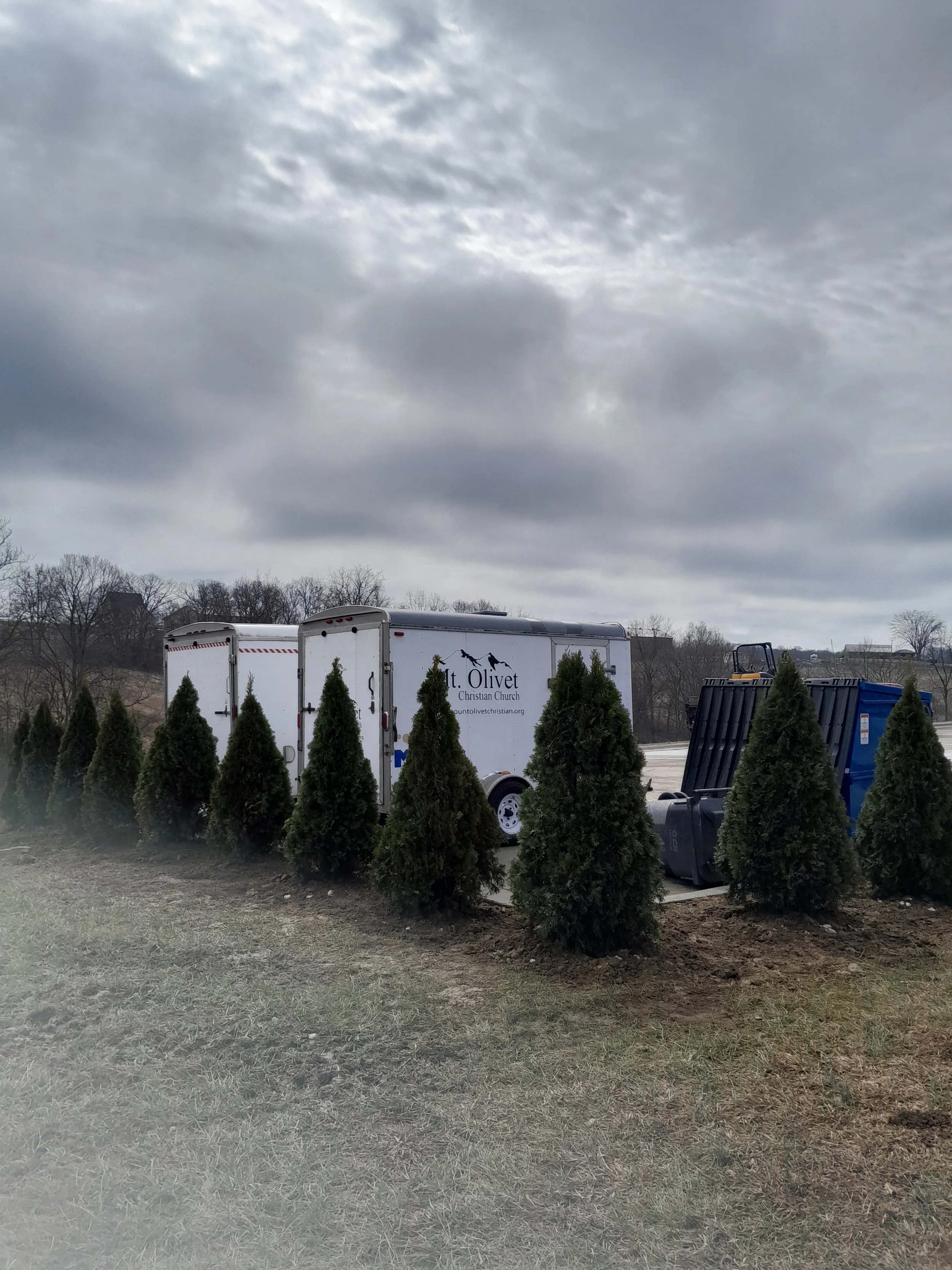 A row of small green trees in front of a white trailer with the logo for Mt. Olivet Christian Church, parked on a street under a cloudy sky.