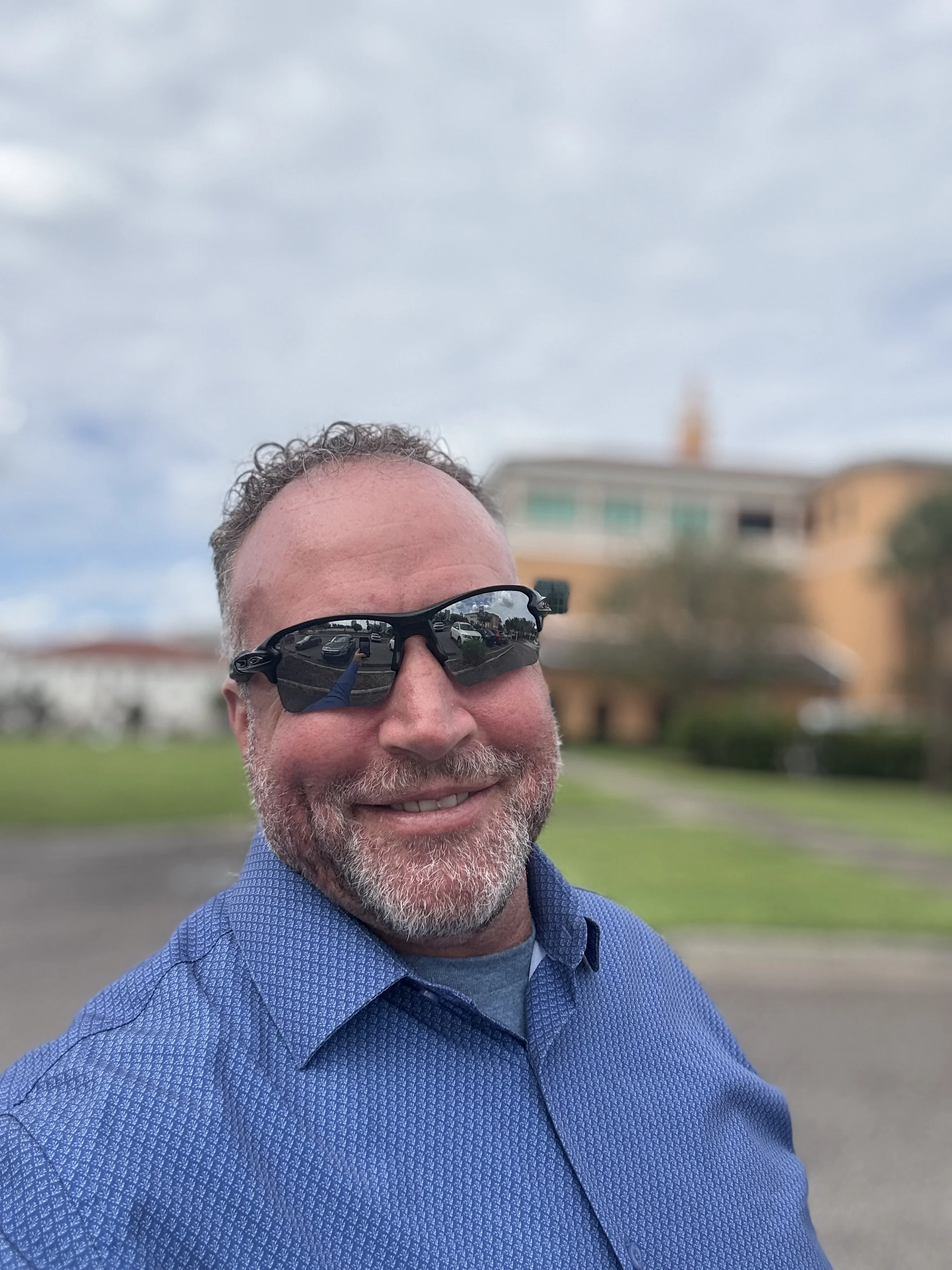 A smiling man with sunglasses and a beard standing outdoors in front of a blurred residential building and a blue sky with some clouds.
Our Licensed Physician, Dustin Finucan.