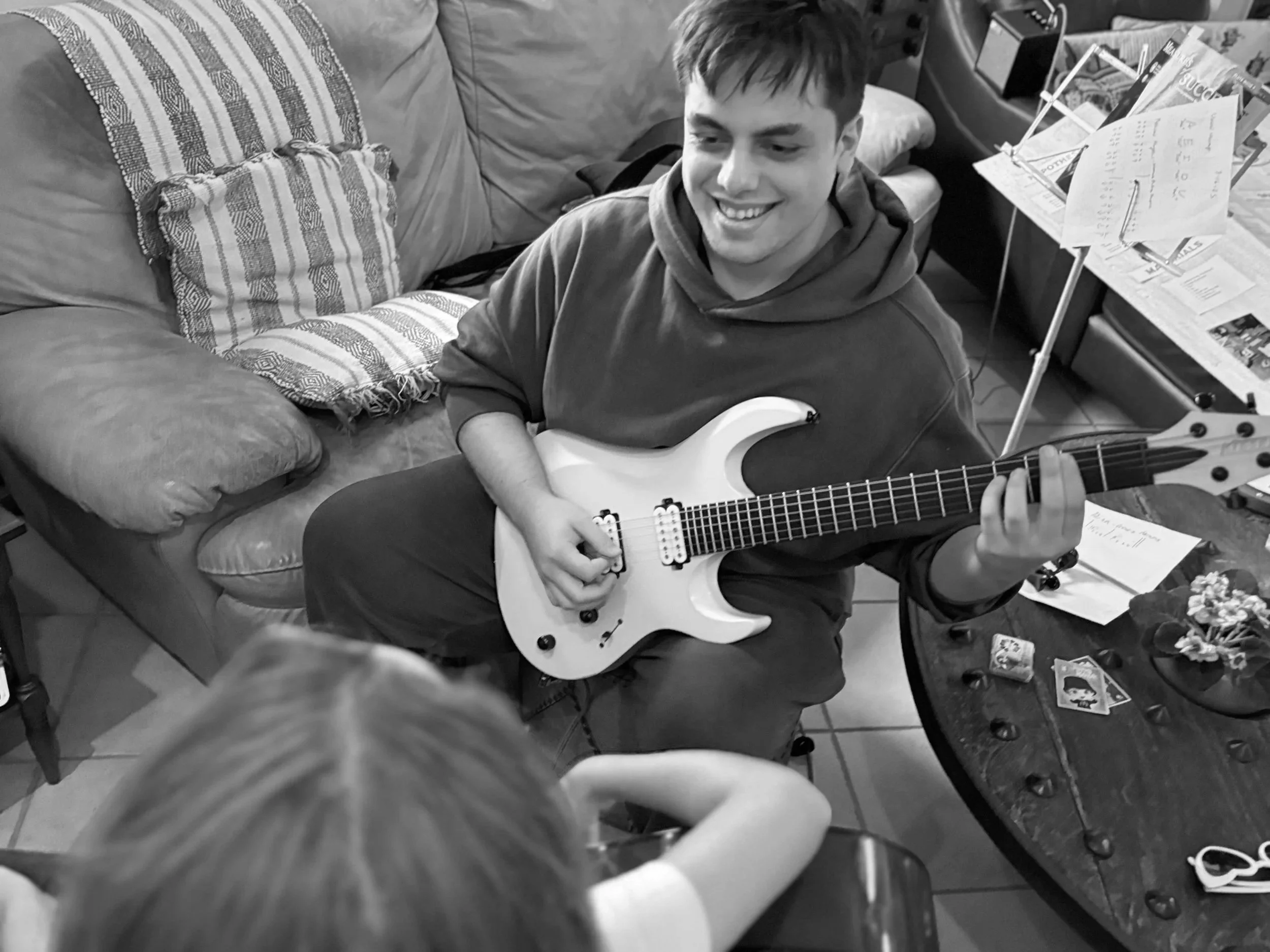 A young man is sitting on a chair playing an electric guitar, smiling. He is in a living room with a sofa, pillows, and a table with scattered papers, cards, and small objects. Someone with blonde hair is partially visible in the foreground.