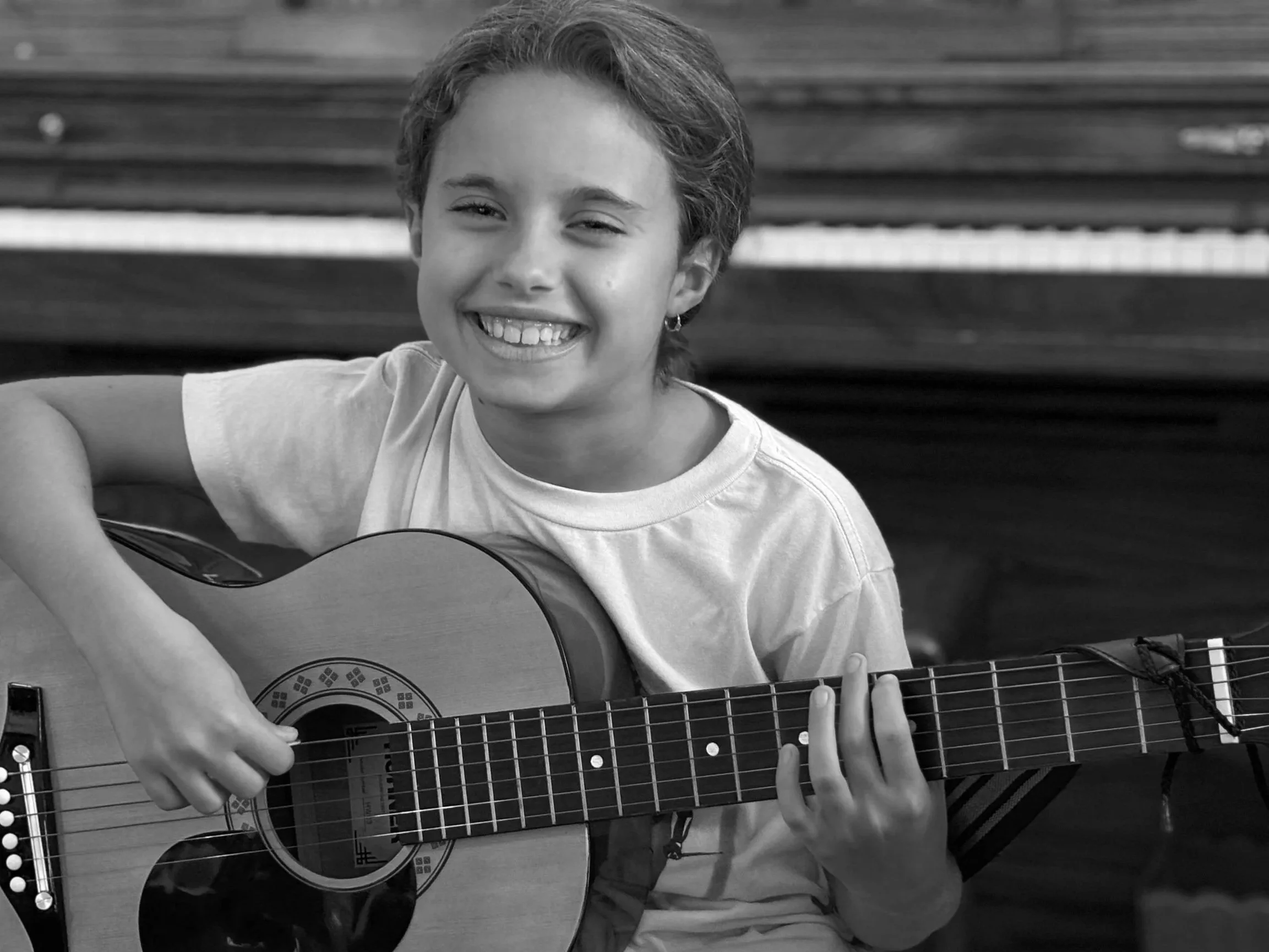 A young girl with short hair smiling and playing an acoustic guitar in a room with a piano in the background.