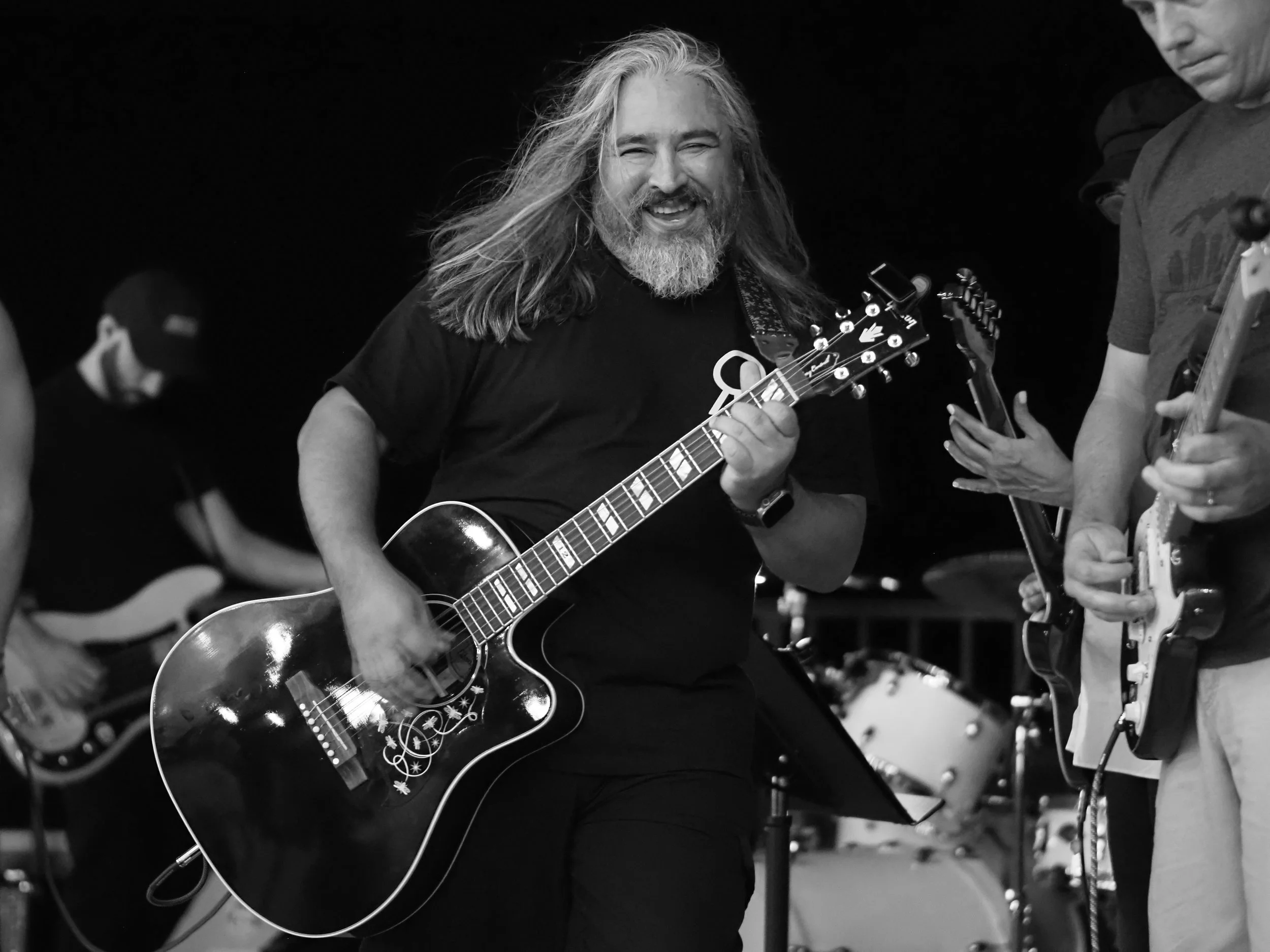 A man with long hair and a beard playing an acoustic guitar, smiling, surrounded by other musicians on stage.