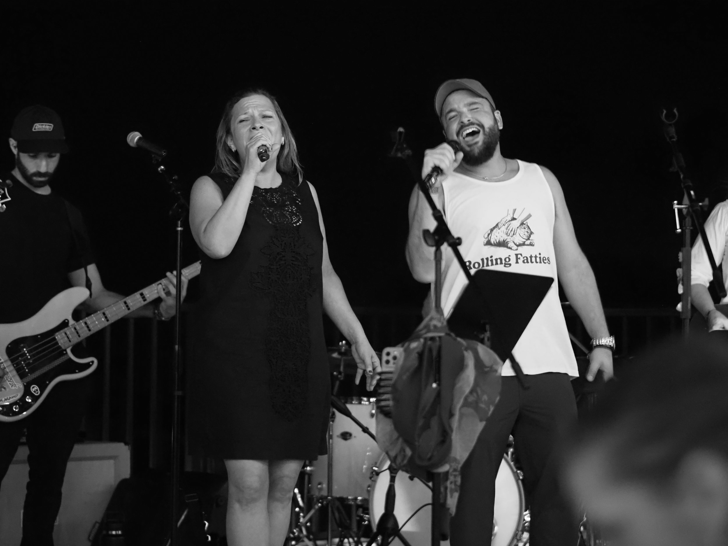 Black and white photo of a band performing on stage with two singers singing into microphones, one woman and one man, and musicians in the background playing instruments.