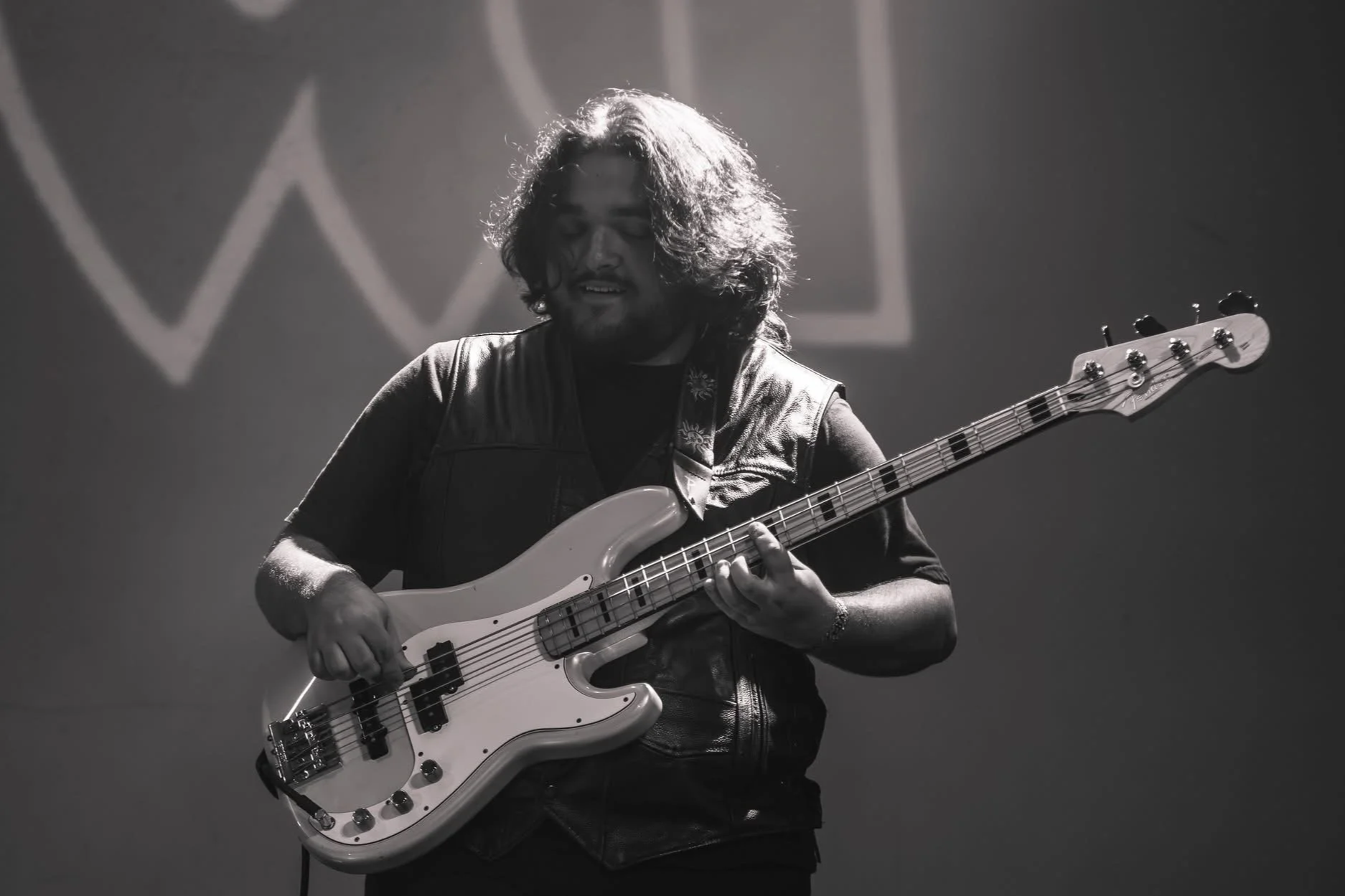 A man with long hair and a beard playing an electric bass guitar on stage, with a dim background and stage lighting.