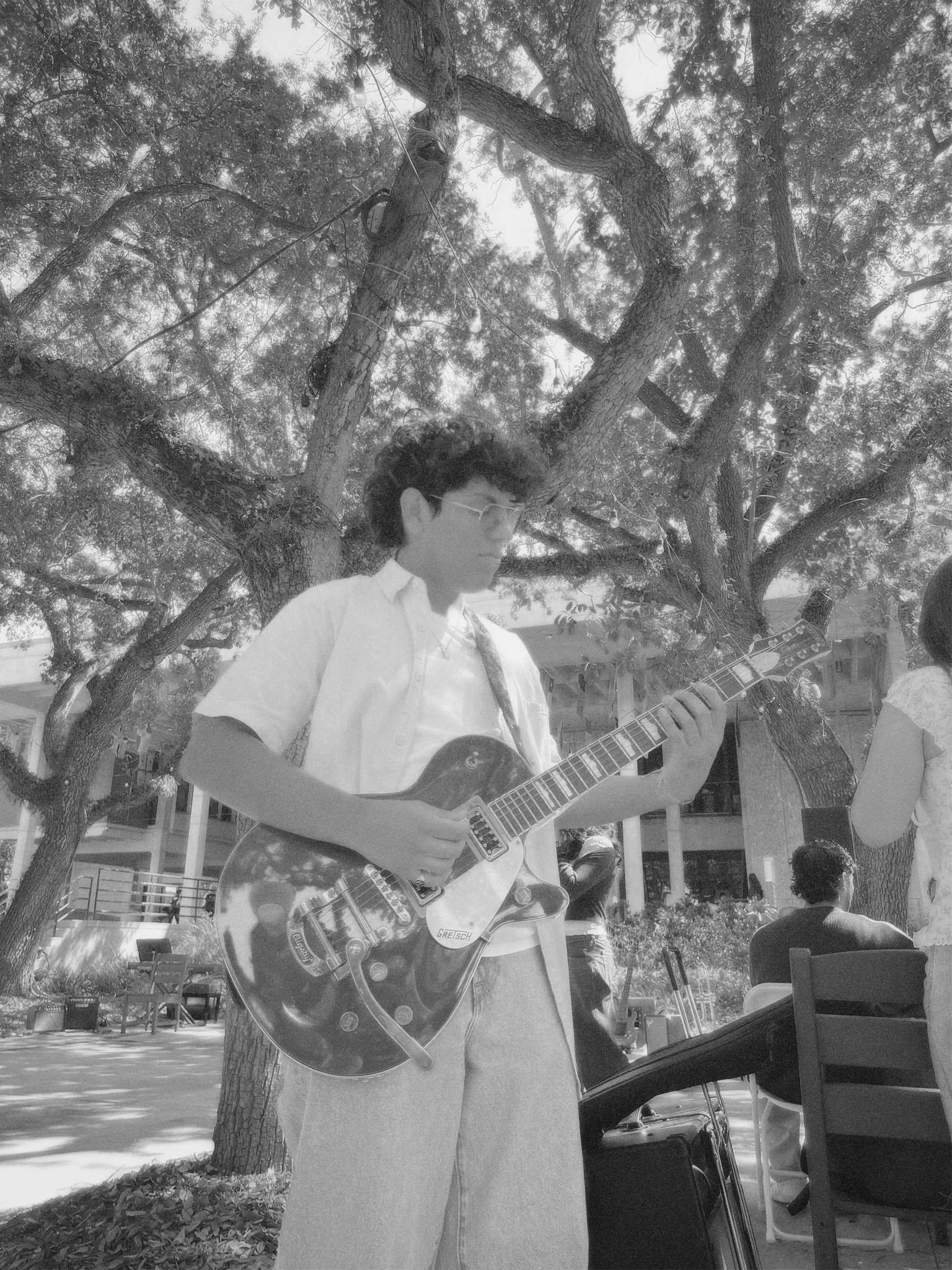 A young man playing an electric guitar outdoors near a tree, with other people around, possibly at a gathering or event.